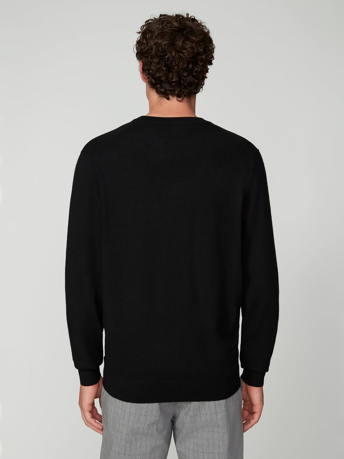A man with curly brown hair stands with his back to the camera, wearing a PROJEK RAW MENS CREW NECK CASHMERE TOUCH SWEATER in black and light gray plaid pants against a plain light background.