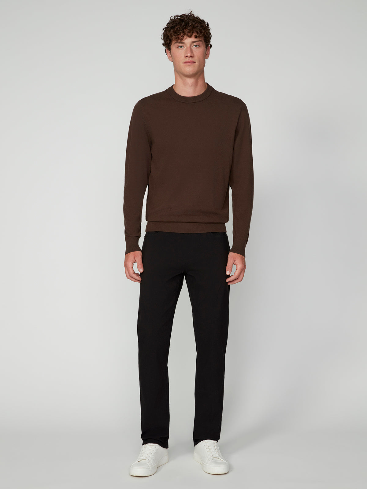 A young man with curly hair faces forward, wearing the PROJEK RAW MENS CREW NECK CASHMERE TOUCH SWEATER in brown, black pants, and white sneakers against a plain light gray background.