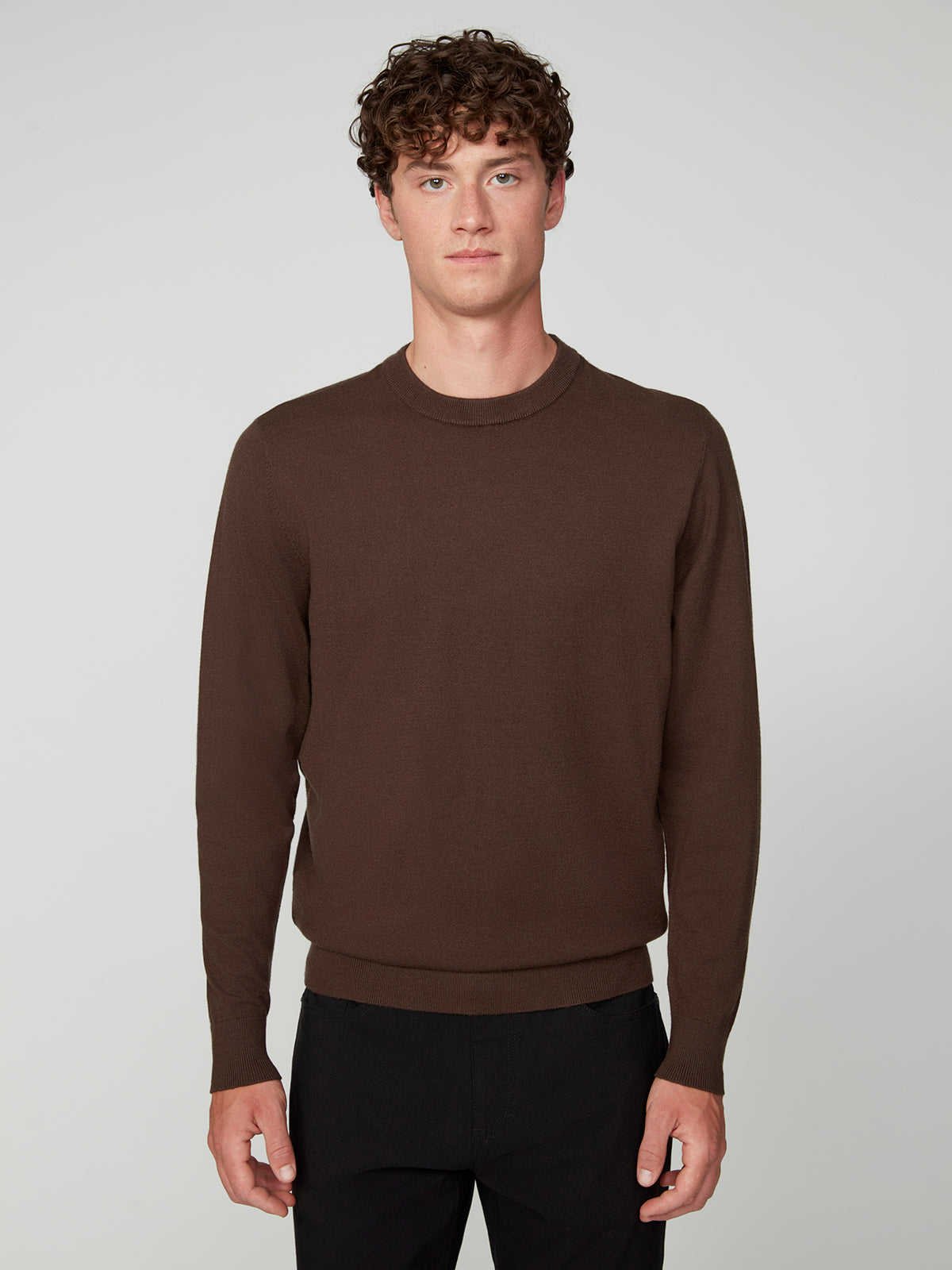 A young man with curly brown hair wears the PROJEK RAW MENS CREW NECK CASHMERE TOUCH SWEATER and black pants, standing against a plain light gray background and looking directly at the camera with a neutral expression.