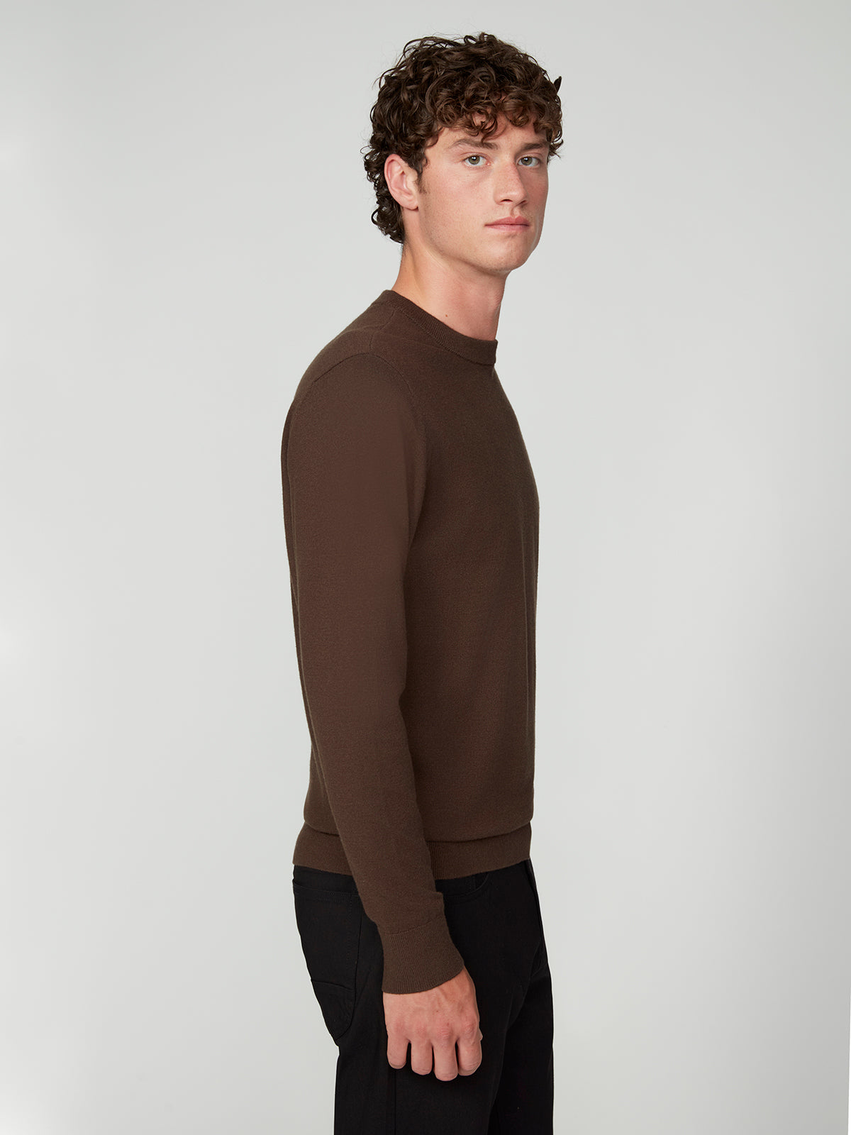 A young man with curly brown hair stands against a light gray background, wearing a PROJEK RAW MENS CREW NECK CASHMERE TOUCH SWEATER in brown and black pants, facing sideways with a neutral expression.