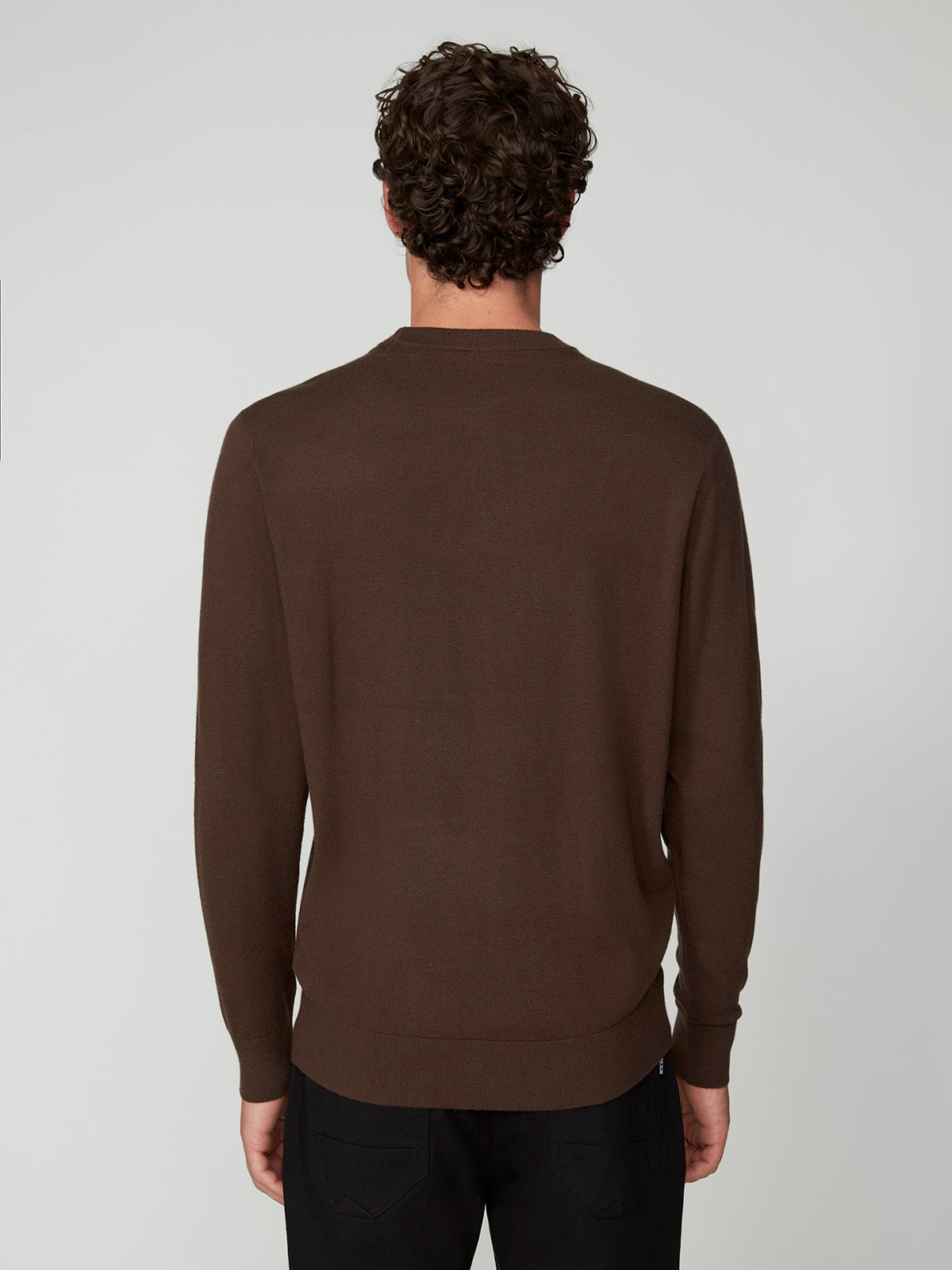 A man with short, curly brown hair is seen from behind, wearing a dark brown PROJEK RAW MENS CREW NECK CASHMERE TOUCH SWEATER and black pants against a plain light gray background.