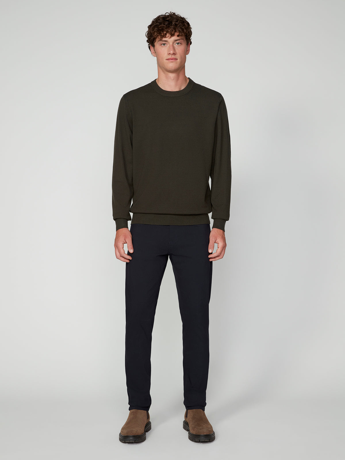 A young man with curly brown hair faces forward, wearing a PROJEK RAW MENS CREW NECK CASHMERE TOUCH SWEATER in dark green, paired with black pants and brown suede shoes, set against a plain light gray background.