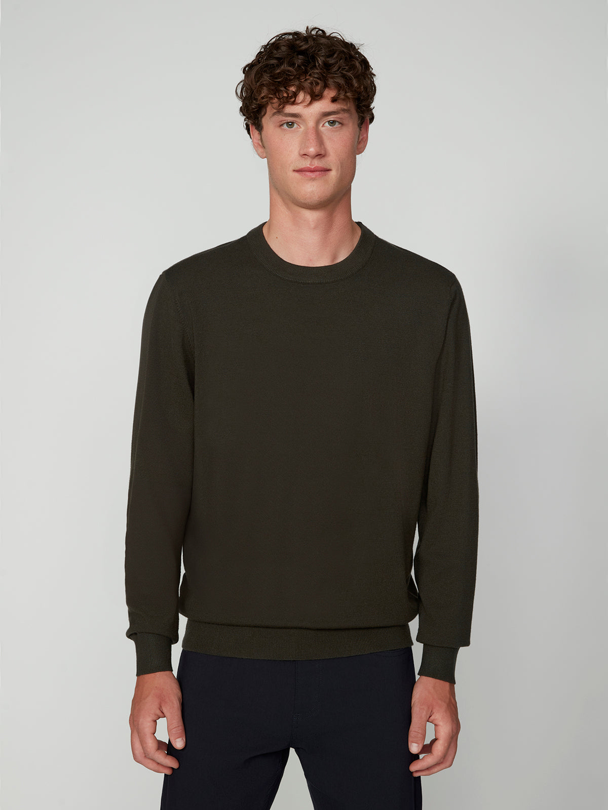 A young man with curly brown hair wears a PROJEK RAW MENS CREW NECK CASHMERE TOUCH SWEATER in dark olive green and dark pants, standing against a light gray background and facing the camera with a neutral expression.