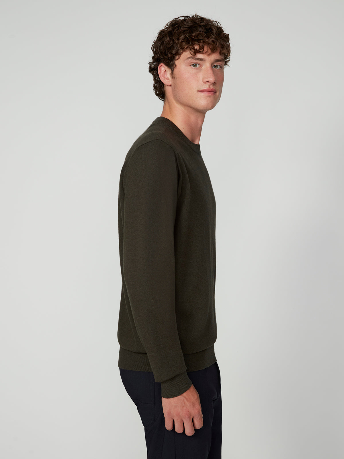 A young man with curly brown hair stands sideways, looking at the camera. He wears a PROJEK RAW MENS CREW NECK CASHMERE TOUCH SWEATER in dark green with dark pants, set against a plain light gray background.