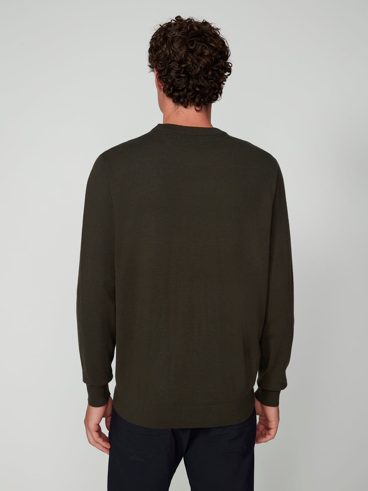 A man with curly brown hair stands facing away from the camera, wearing a PROJEK RAW MENS CREW NECK CASHMERE TOUCH SWEATER and dark pants against a plain light gray background.