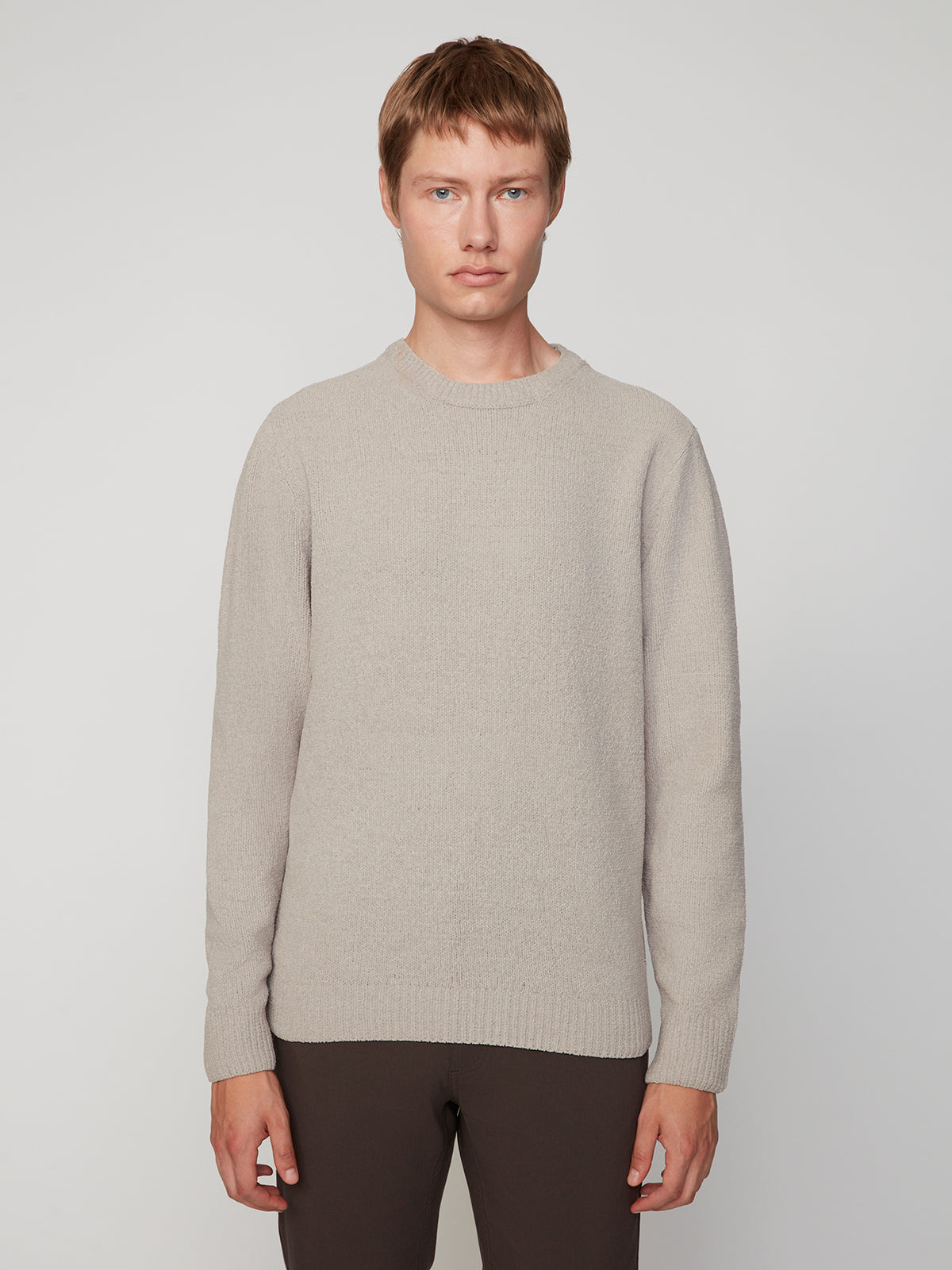 A young person with short light brown hair stands against a plain background, wearing the PROJEK RAW MENS CREW NECK CHENILLE SWEATER in beige and dark pants, facing the camera with a neutral expression.