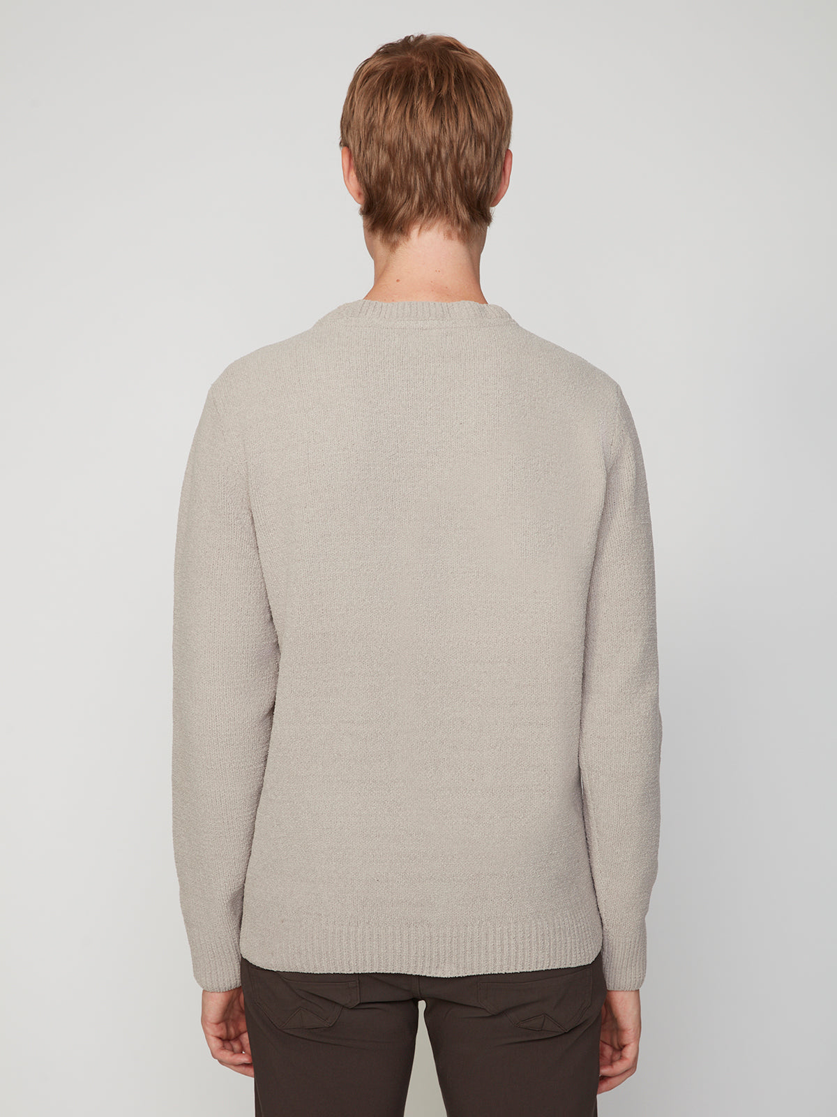 A person with short light brown hair stands facing away from the camera, wearing the PROJEK RAW MENS CREW NECK CHENILLE SWEATER in light beige and dark pants against a plain white background.