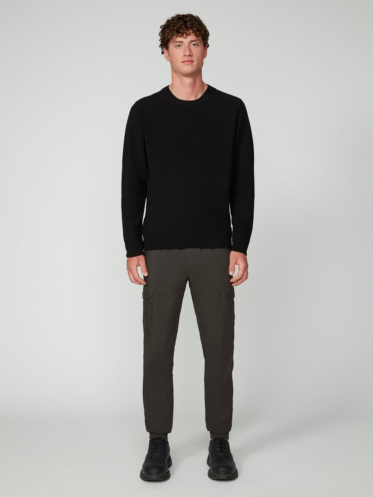 A young man with short curly hair stands facing forward, wearing the PROJEK RAW MENS CREW NECK CHENILLE SWEATER in black, paired with olive green cargo pants and black boots, set against a plain light gray background.
