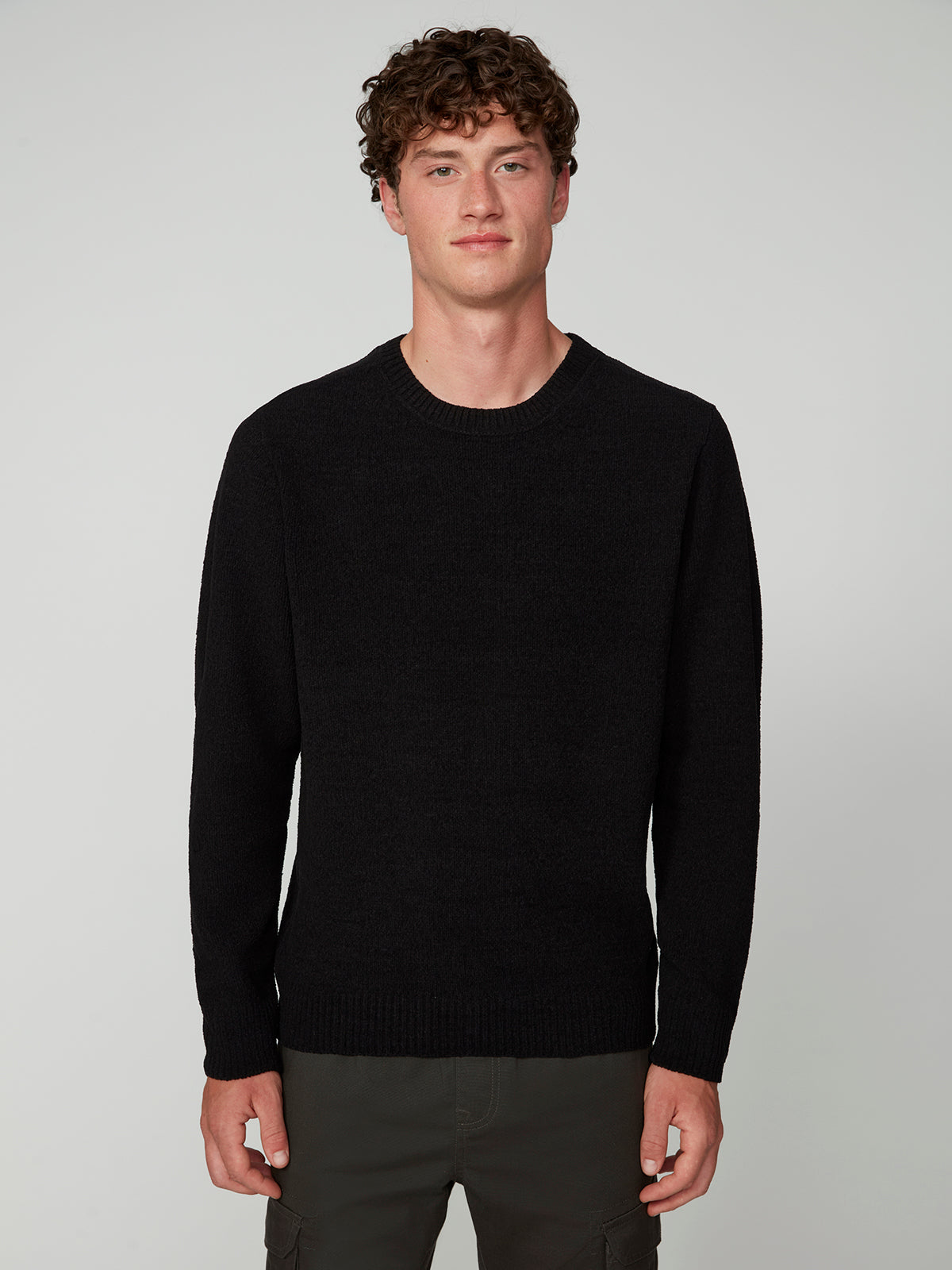 A young man with curly brown hair wears the PROJEK RAW MENS CREW NECK CHENILLE SWEATER in black and dark pants, standing against a light gray background, facing forward with a neutral expression.