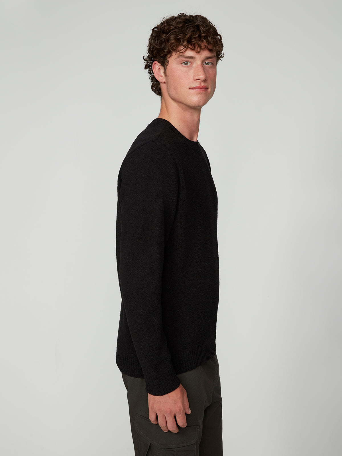 A young man with curly brown hair stands sideways, facing left, wearing a PROJEK RAW MENS CREW NECK CHENILLE SWEATER in black with olive green pants against a light gray background.