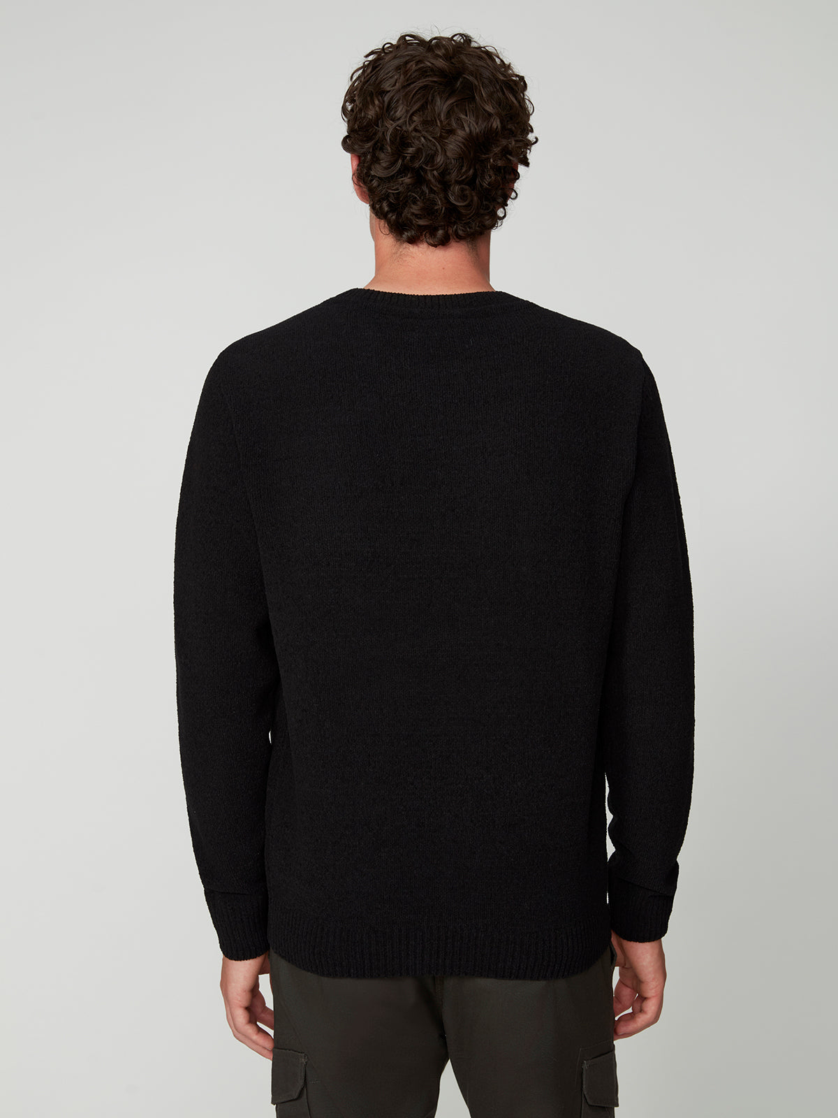 A man with curly hair, seen from behind, wears a PROJEK RAW MENS CREW NECK CHENILLE SWEATER in black and dark pants against a neutral background.