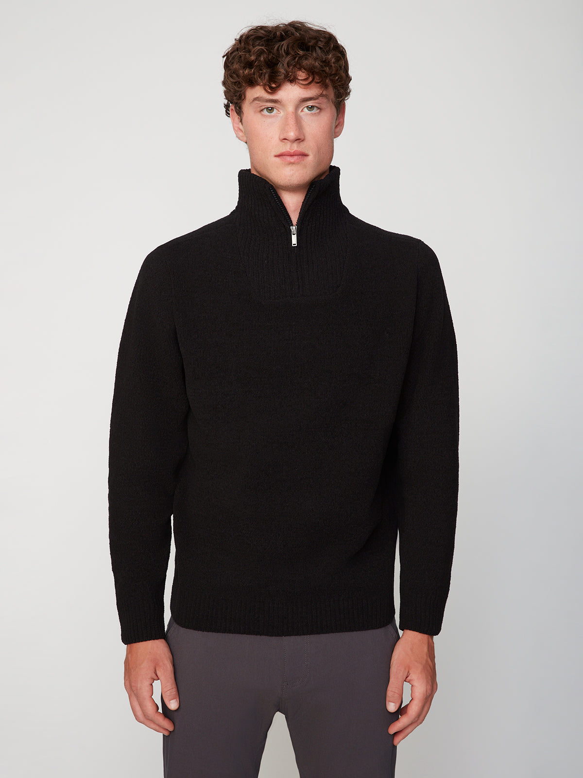 A young man with curly hair wears the PROJEK RAW MENS CHENILLE MOCK NECK SWEATER in black and gray pants, standing against a plain light background, facing forward with a neutral expression.