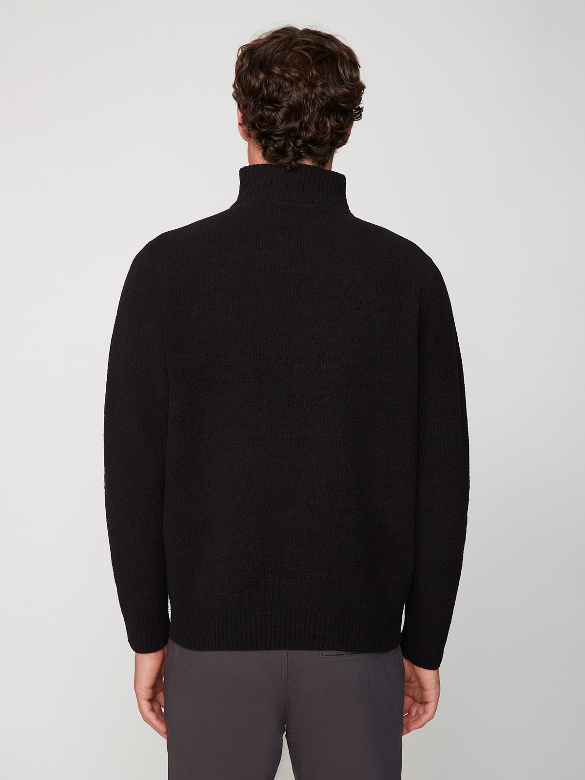 A person with short curly brown hair is seen from the back, wearing a black PROJEK RAW MENS CHENILLE MOCK NECK SWEATER and gray pants against a plain light background.