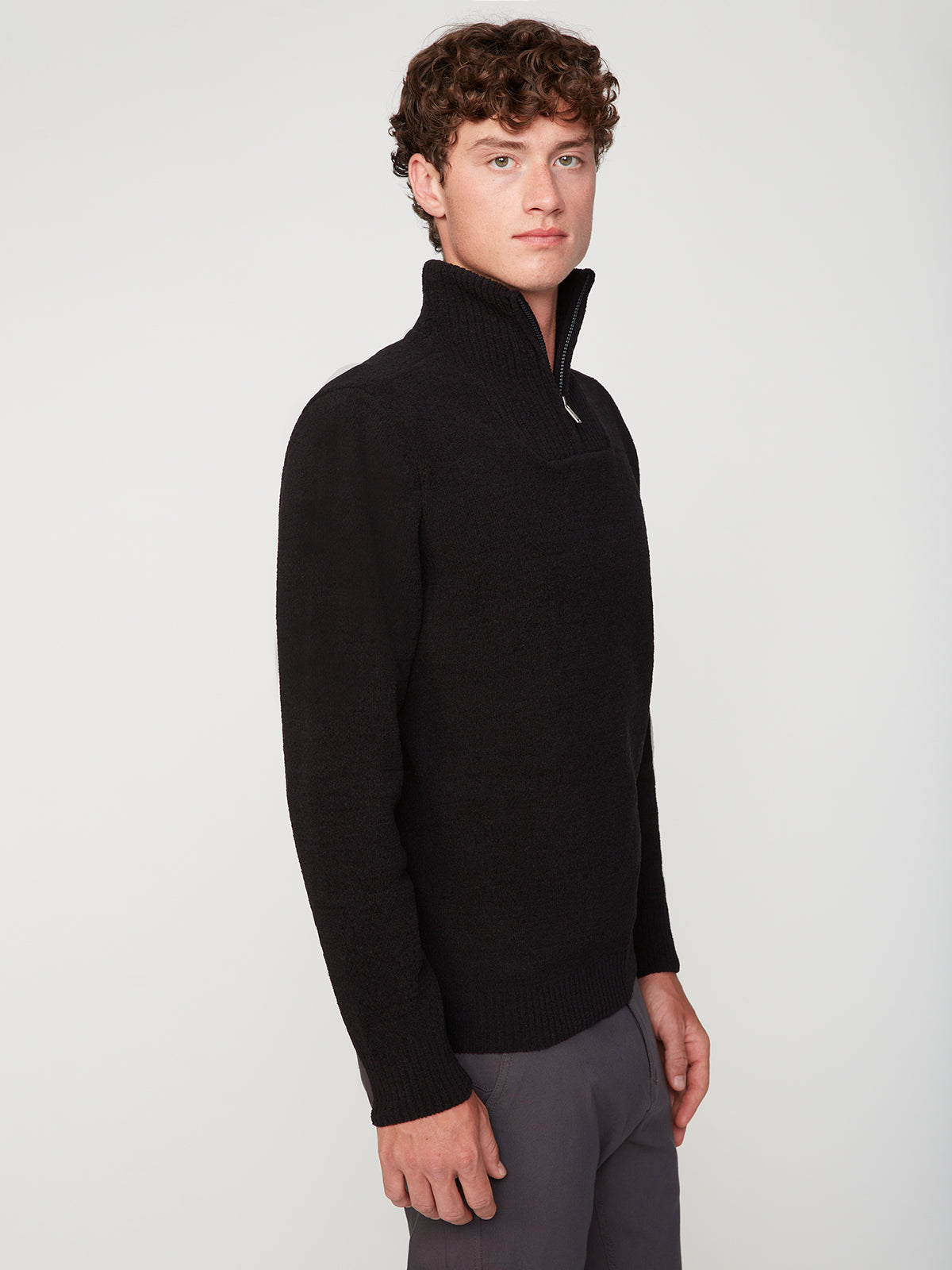 A young man with curly brown hair models the PROJEK RAW MENS CHENILLE MOCK NECK SWEATER in black with gray pants, standing sideways and looking at the camera against a plain white background.
