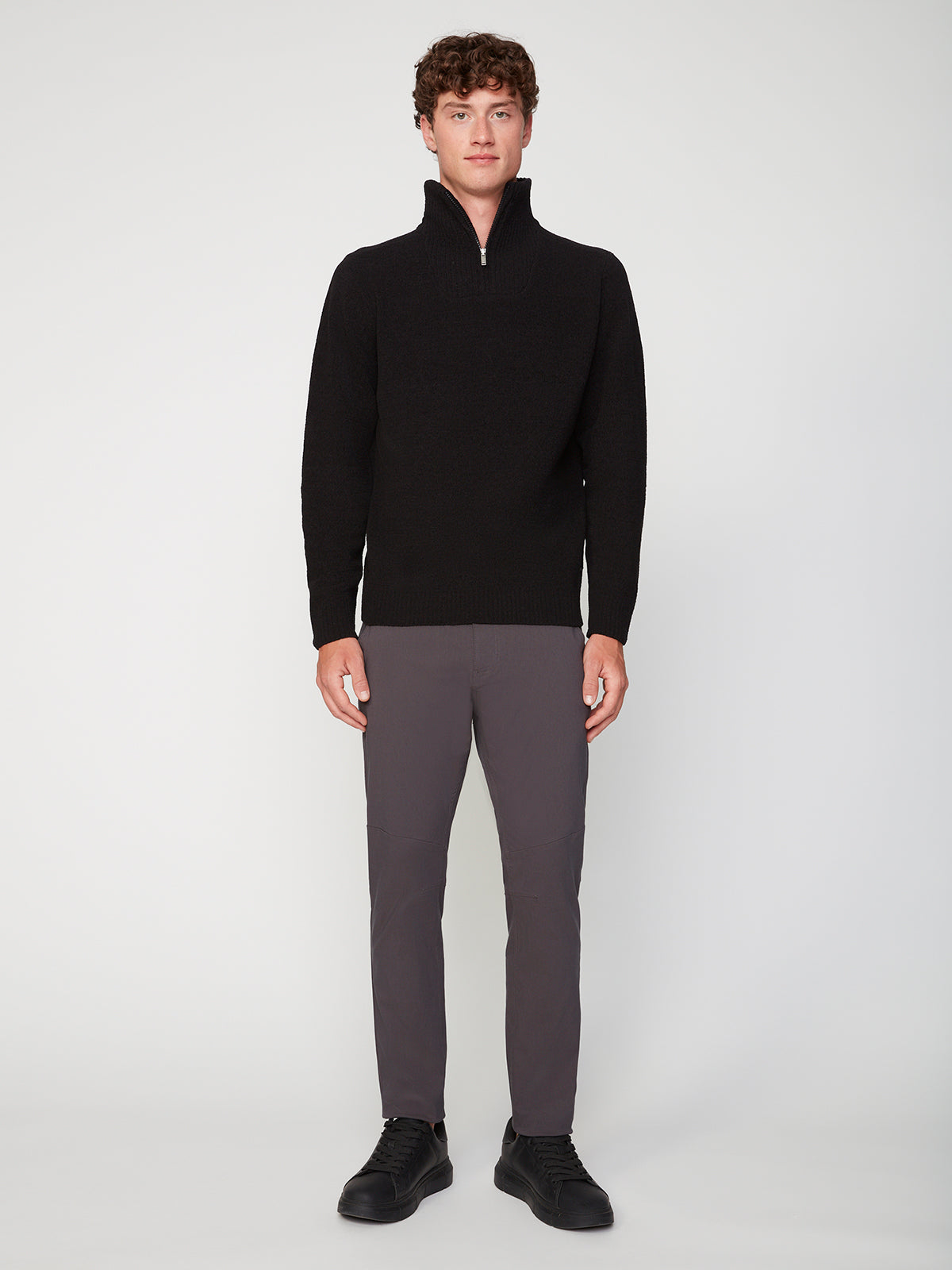 A young man with short curly hair and a neutral expression stands against a plain background, wearing the PROJEK RAW MENS CHENILLE MOCK NECK SWEATER in black, paired with gray slim-fit pants and black shoes.
