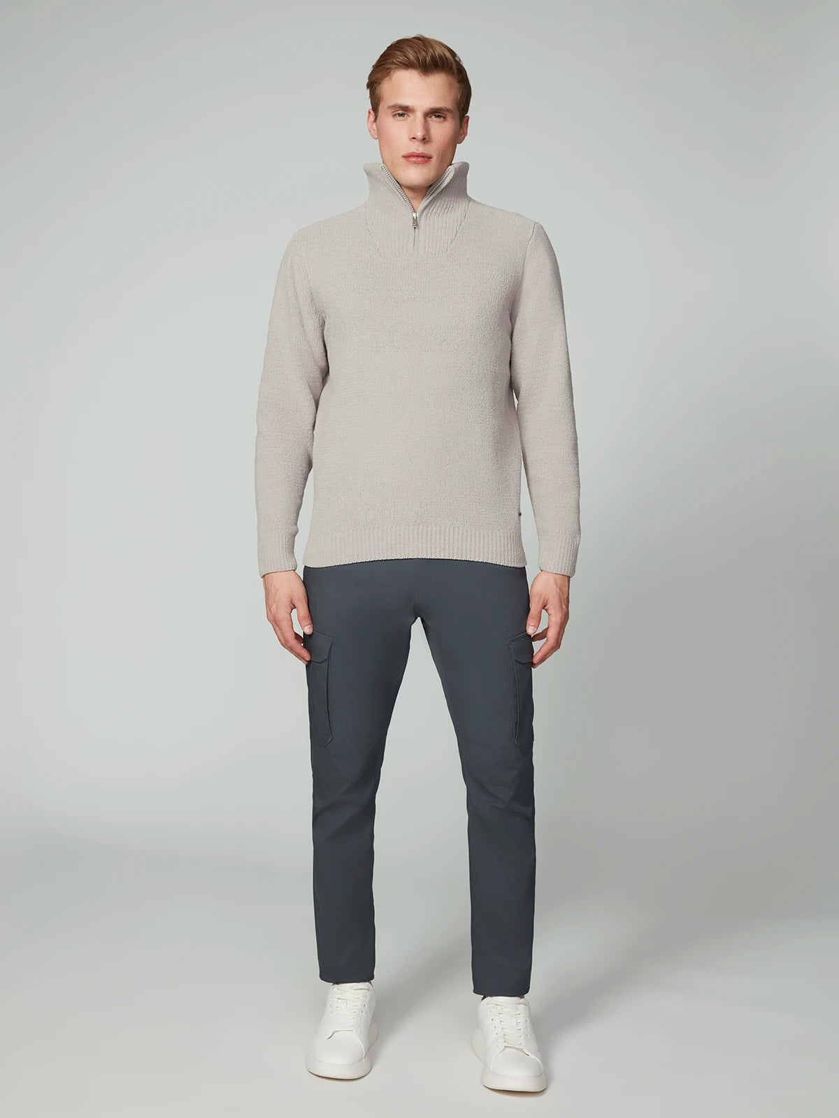 A person stands facing forward in a light beige PROJEK RAW MENS CHENILLE MOCK NECK SWEATER, dark gray cargo pants, and white sneakers against a plain light gray background.