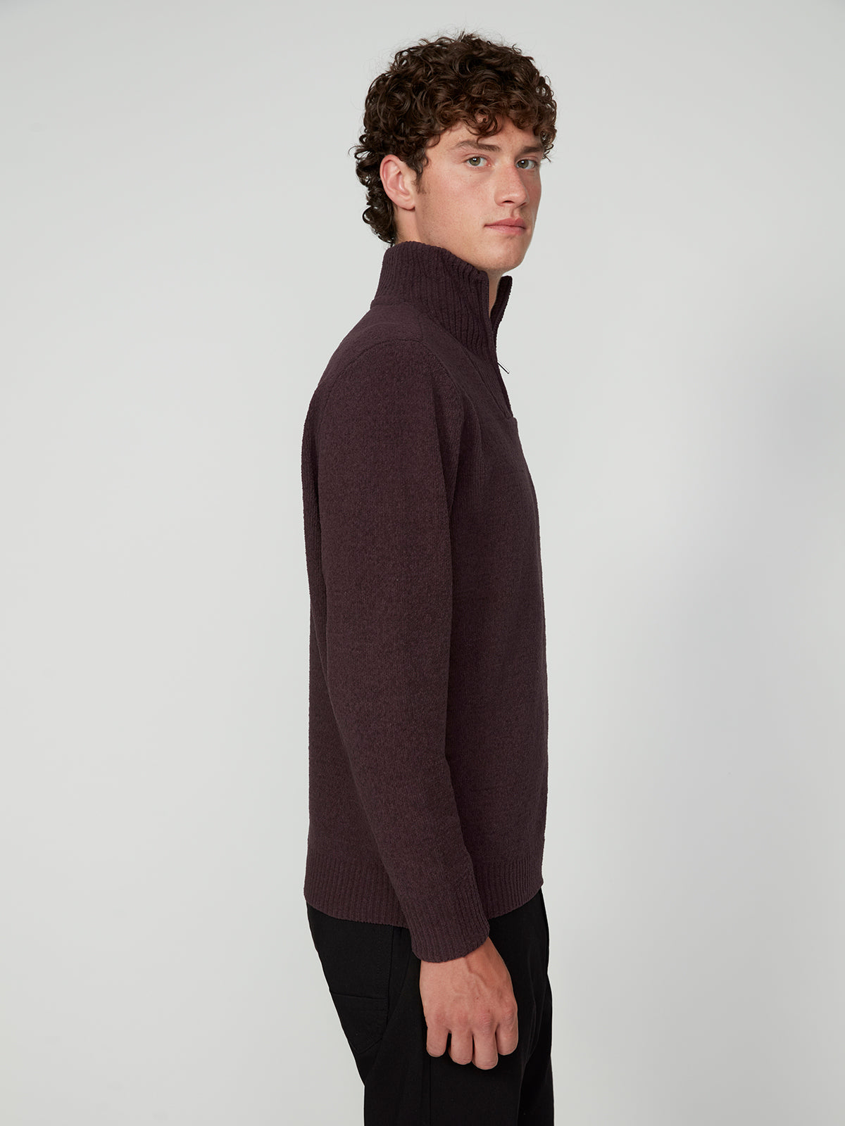 A young man with short curly hair stands sideways against a plain light background, wearing black pants and a PROJEK RAW MENS CHENILLE MOCK NECK SWEATER in dark purple.