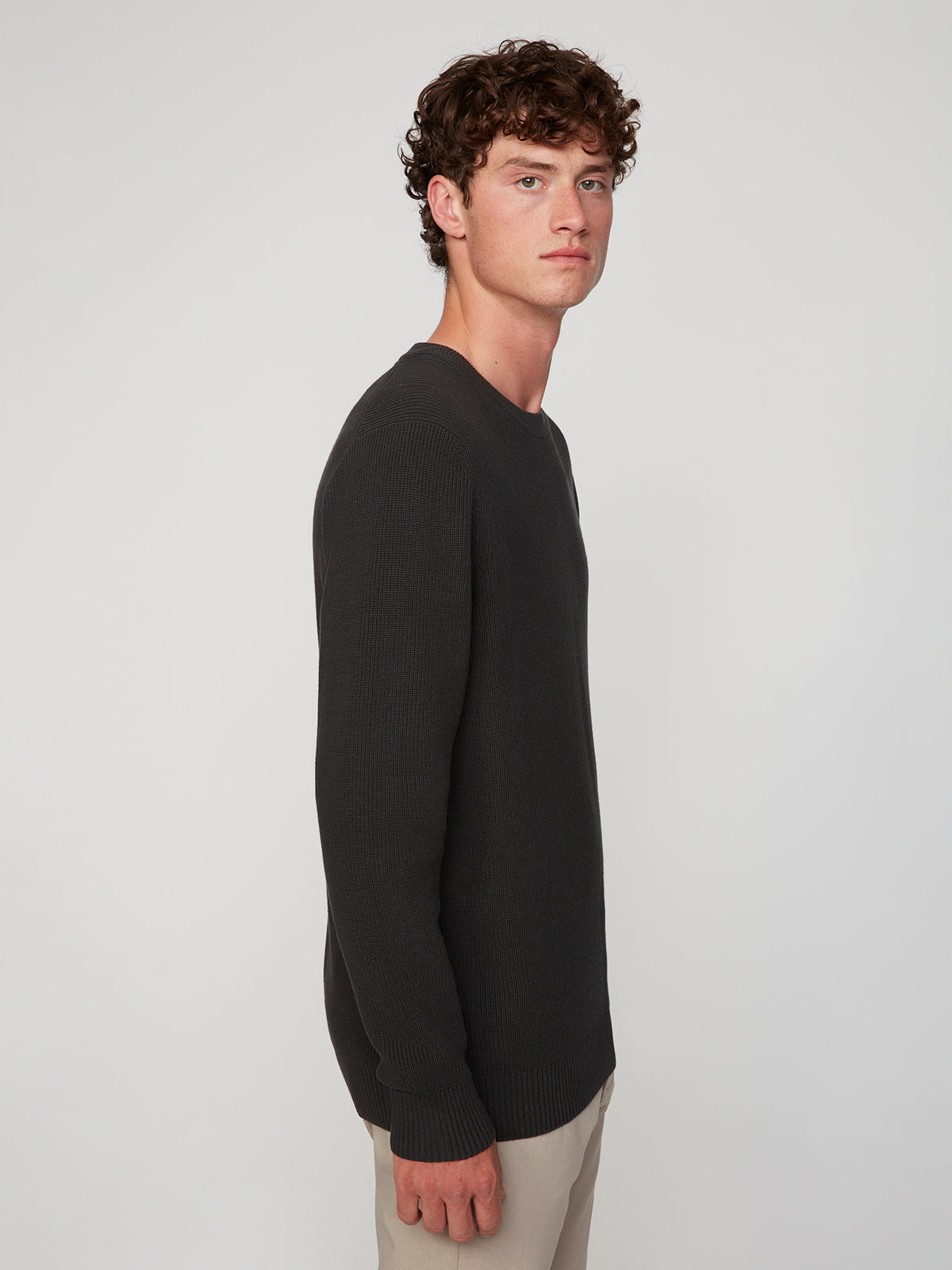 A young man with curly brown hair wears a PROJEK RAW MENS SHAKER RIB KNIT SWEATER in a dark shade and light pants, standing sideways and looking toward the camera against a plain light background.