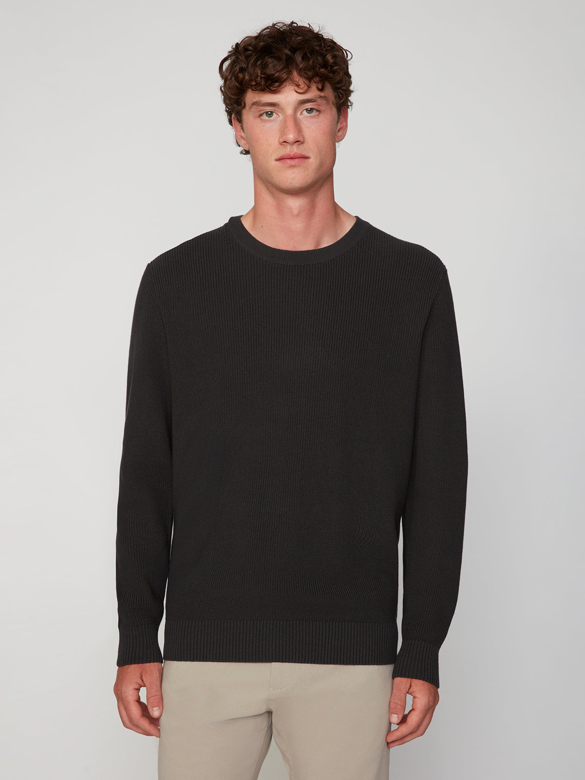 A young man with curly brown hair wears the PROJEK RAW MENS SHAKER RIB KNIT SWEATER in black and light beige pants, standing against a plain light gray background, facing forward with a neutral expression.