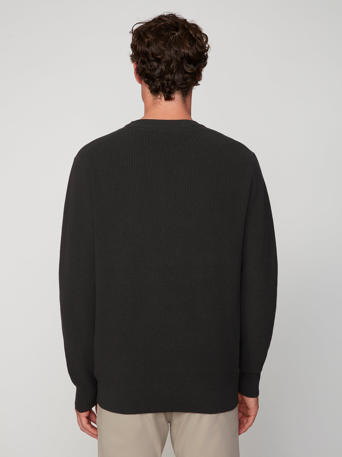 A man with short, curly brown hair stands with his back to the camera, wearing the PROJEK RAW MENS SHAKER RIB KNIT SWEATER and light beige pants against a plain light gray background.