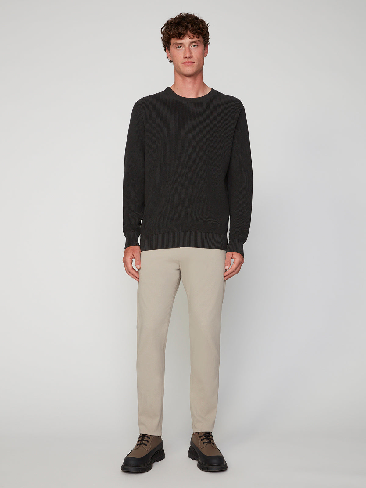 A young man faces forward against a light gray background, wearing the PROJEK RAW MENS SHAKER RIB KNIT SWEATER in black, paired with light beige pants and brown lace-up shoes.
