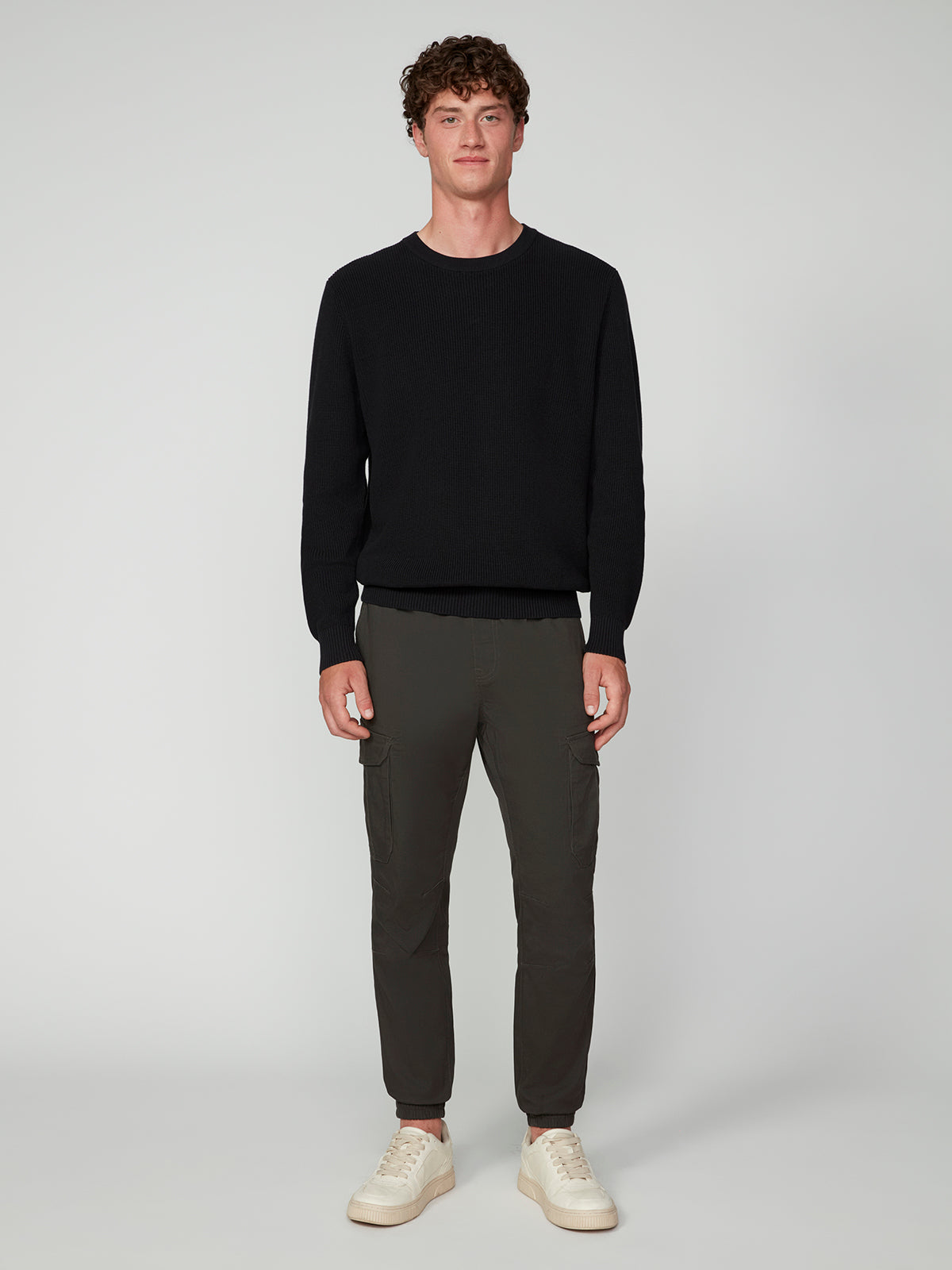 A young man with short curly hair faces forward, wearing a PROJEK RAW MENS SHAKER RIB KNIT SWEATER, dark gray cargo pants, and white sneakers against a plain light gray background.