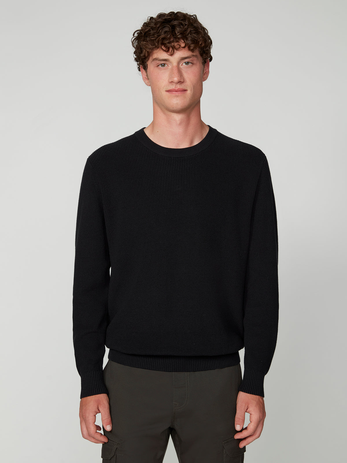 A young man with curly brown hair stands facing forward, wearing a PROJEK RAW MENS SHAKER RIB KNIT SWEATER and dark pants against a plain light gray background.