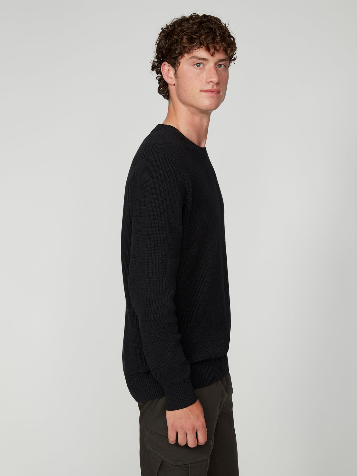A young man with curly brown hair stands sideways, facing left. Hes wearing a PROJEK RAW MENS SHAKER RIB KNIT SWEATER and dark pants, posing against a plain light gray background.