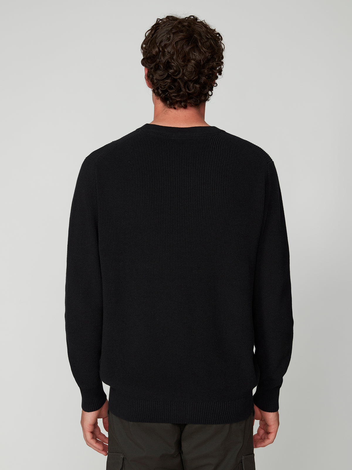 A man with curly brown hair is seen from behind, wearing a black PROJEK RAW MENS SHAKER RIB KNIT SWEATER and dark pants, standing against a plain light gray background.