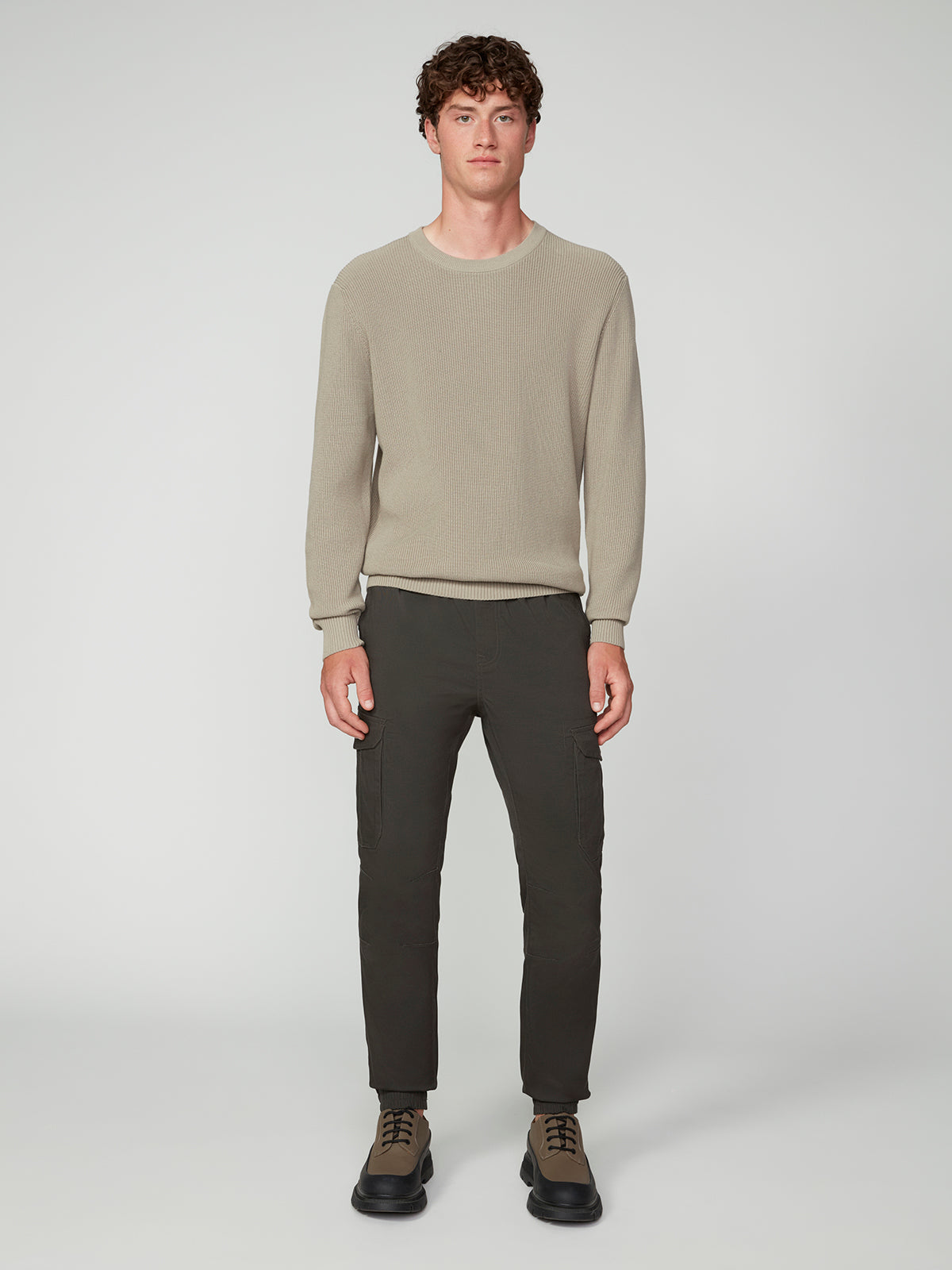 A young man with short curly hair faces forward, wearing a beige PROJEK RAW MENS SHAKER RIB KNIT SWEATER, dark olive cargo pants, and beige lace-up shoes against a plain light gray background.