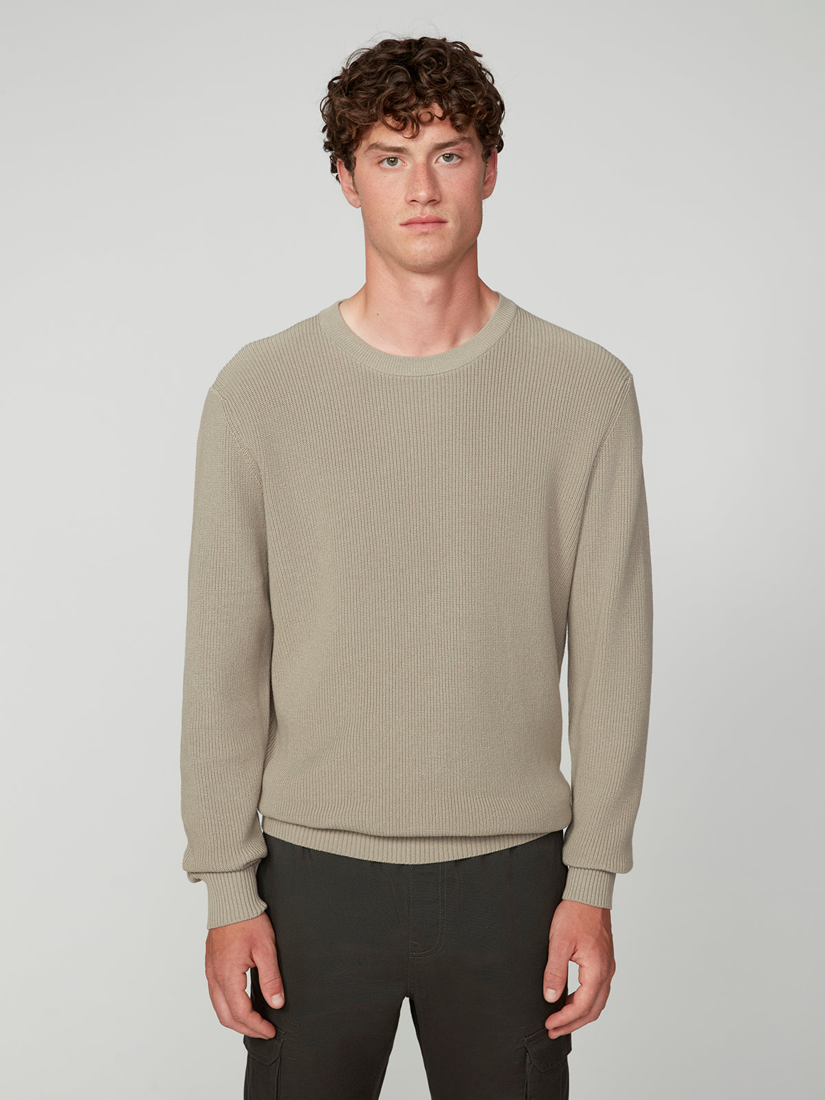 A young man with curly brown hair wears the PROJEK RAW MENS SHAKER RIB KNIT SWEATER in beige and dark pants, standing against a plain light gray background with a neutral expression.