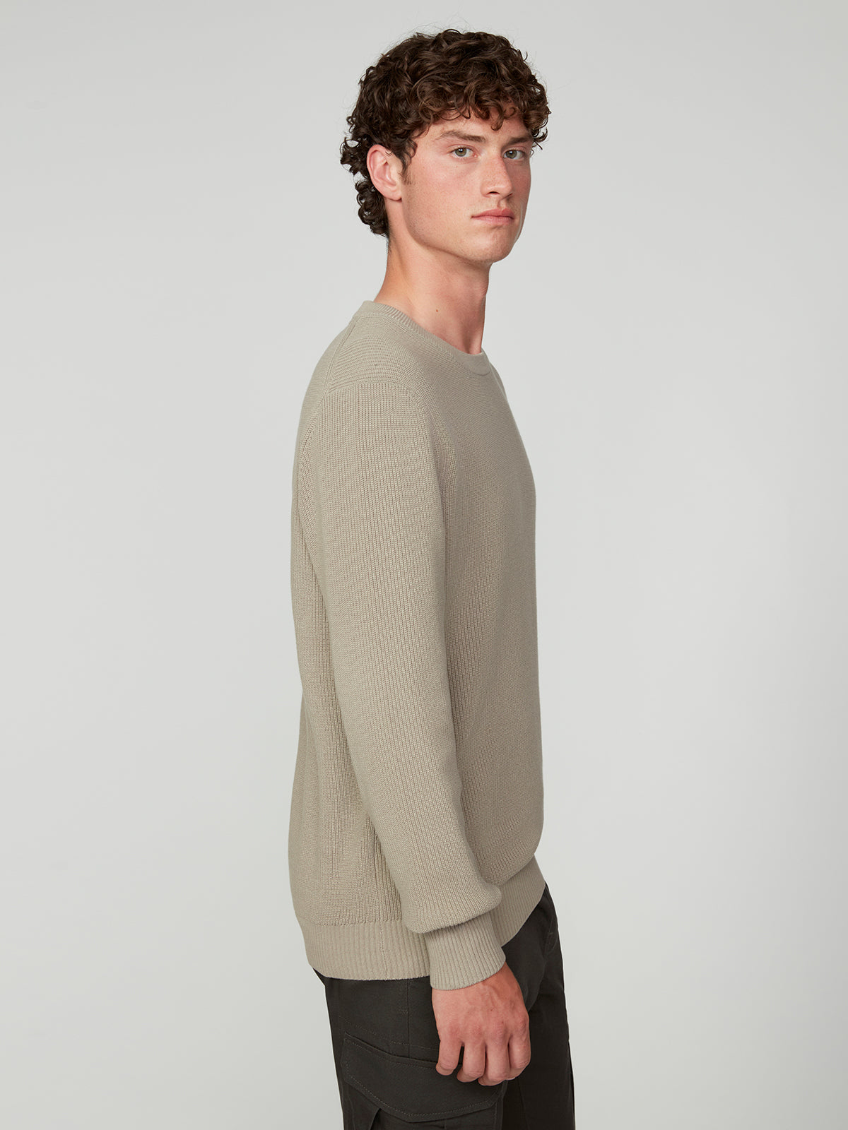 A young man with curly brown hair wears the PROJEK RAW MENS SHAKER RIB KNIT SWEATER in beige and dark pants, standing in profile against a plain light gray background.