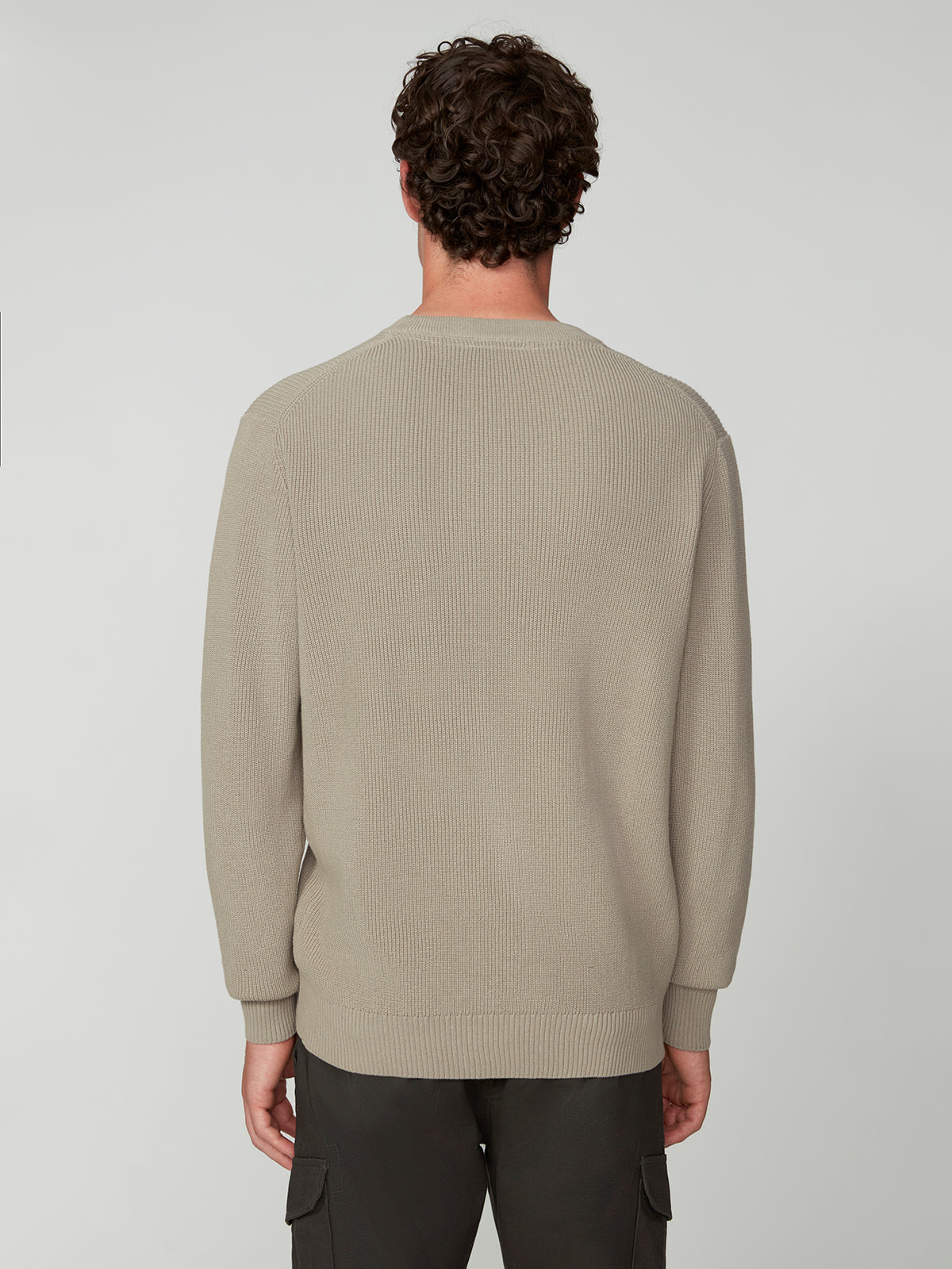 A man with short curly hair, seen from behind, wears the PROJEK RAW MENS SHAKER RIB KNIT SWEATER in light beige and dark pants, standing against a plain light gray background.
