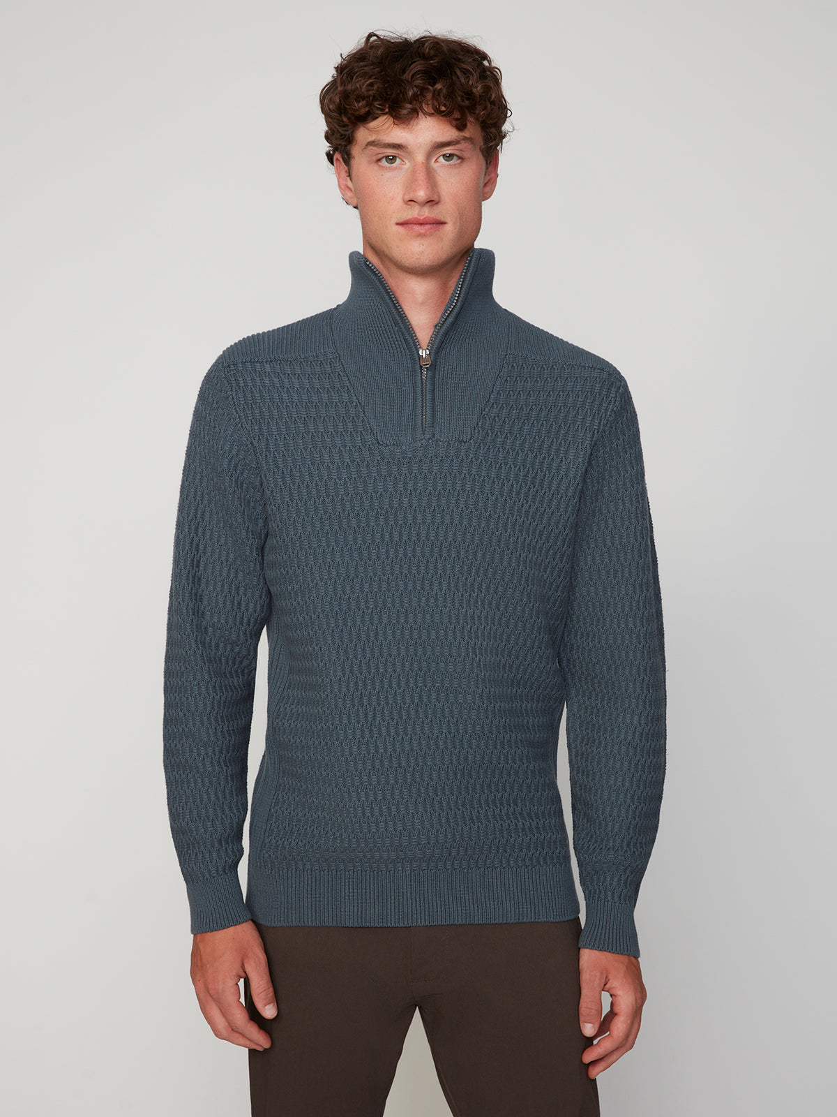 A young man with curly brown hair wears the PROJEK RAW MENS 1/4 ZIP JACQUARD KNIT MOCK NECK SWEATER in blue and brown pants, standing against a plain light gray background.