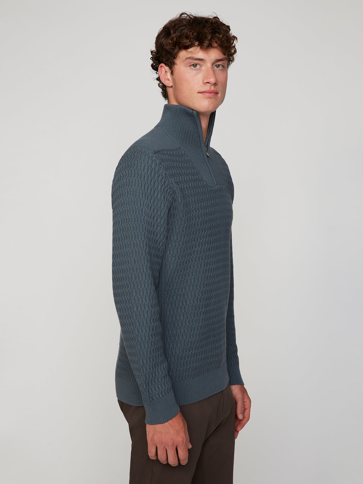 A young man with curly brown hair stands sideways, wearing the PROJEK RAW MENS 1/4 ZIP JACQUARD KNIT MOCK NECK SWEATER and brown pants, looking forward with a neutral expression against a plain light background.