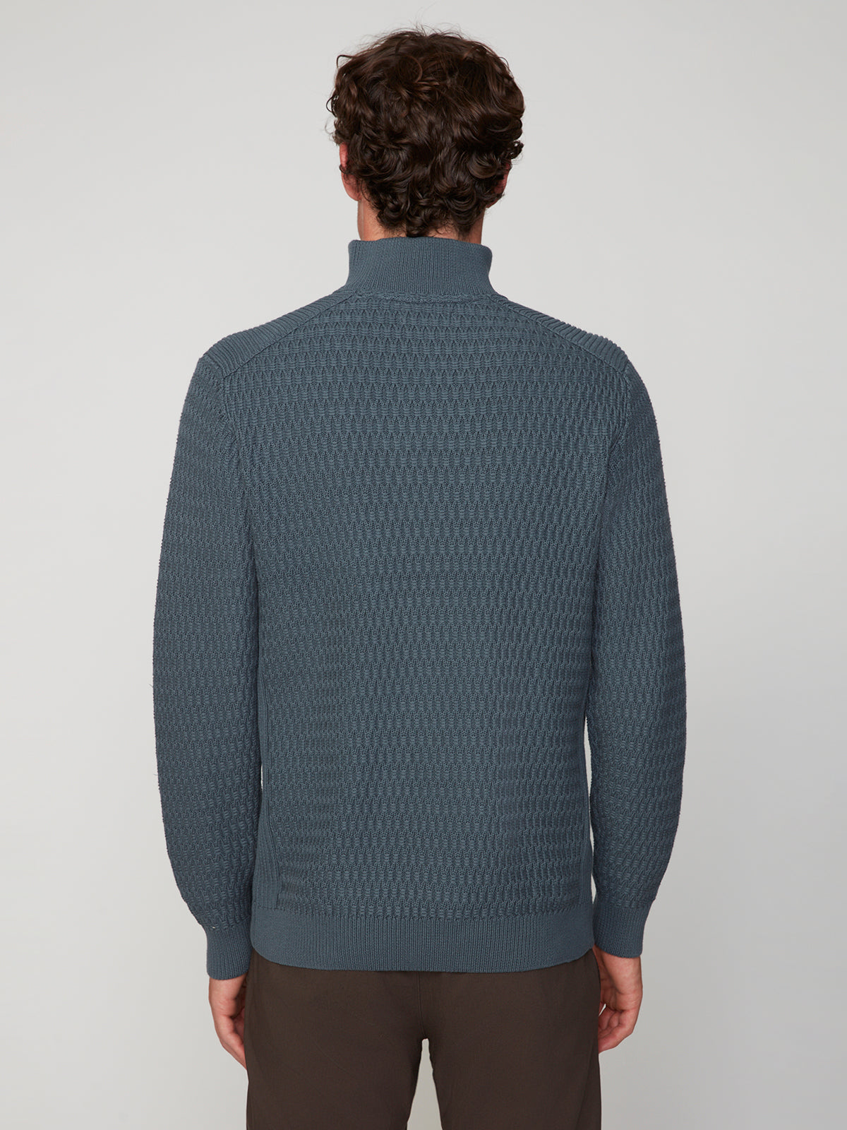 A person with short, curly brown hair is seen from behind, wearing the PROJEK RAW MENS 1/4 ZIP JACQUARD KNIT MOCK NECK SWEATER in blue with brown pants against a plain light gray background.