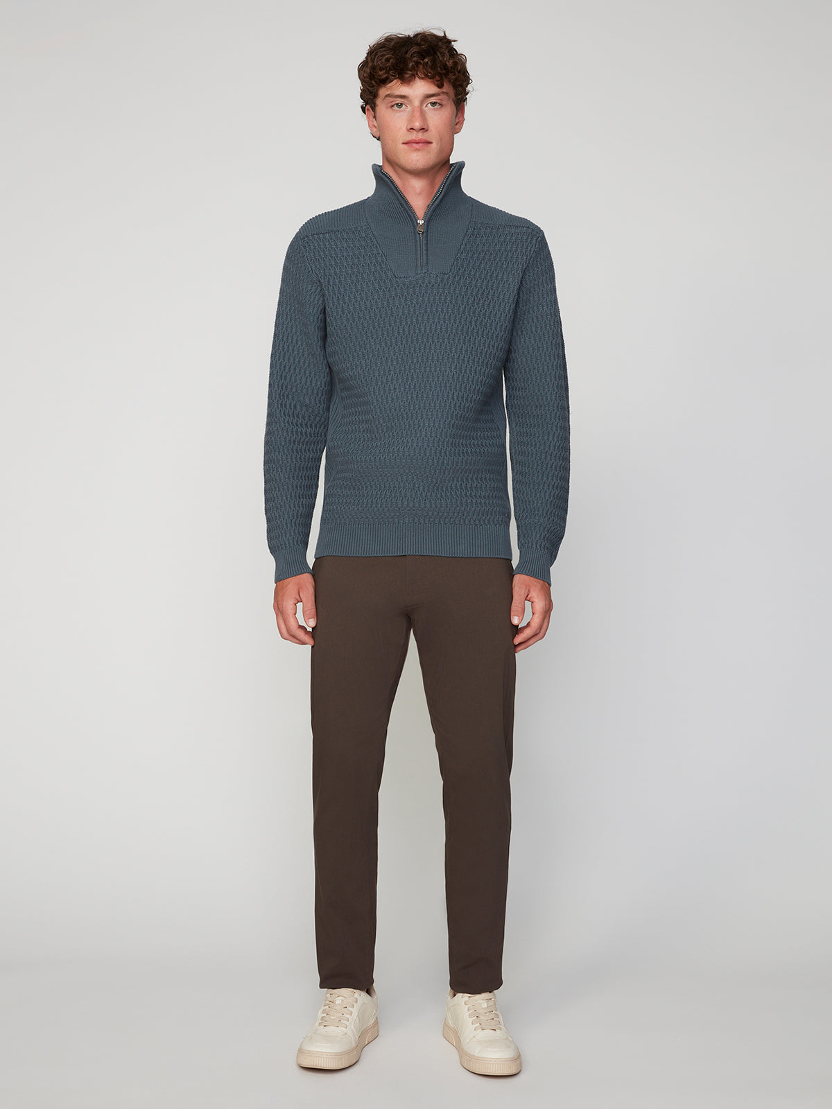 A man faces the camera in a PROJEK RAW MENS 1/4 ZIP JACQUARD KNIT MOCK NECK SWEATER, dark brown pants, and white sneakers against a plain light gray background.
