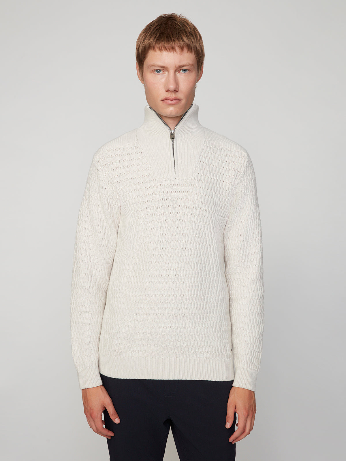 A man with short light brown hair models the PROJEK RAW MENS 1/4 ZIP JACQUARD KNIT MOCK NECK SWEATER in cream and dark pants, standing against a light gray background and facing the camera with a neutral expression.