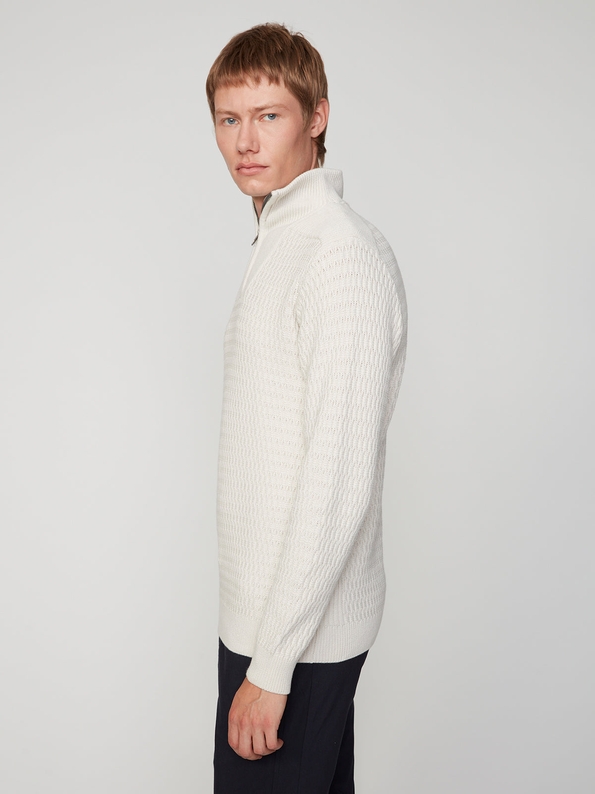 A young man with short light brown hair models the PROJEK RAW MENS 1/4 ZIP JACQUARD KNIT MOCK NECK SWEATER and black pants, standing sideways with a neutral expression against a plain light background.