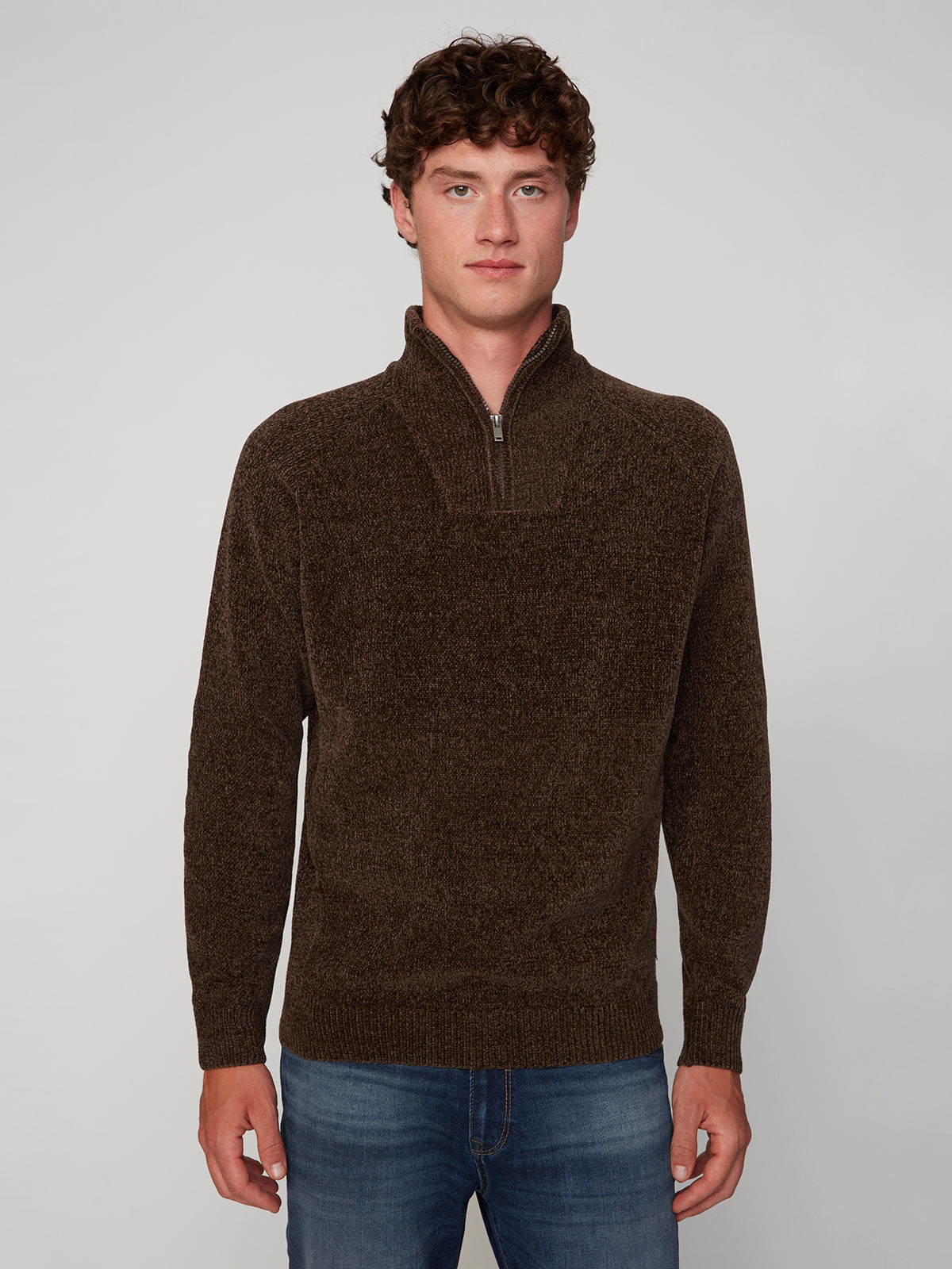 A man with short curly brown hair wears the PROJEK RAW MENS CHENILLE MOCK NECK SWEATER in textured dark brown and blue jeans, standing against a plain light gray background.