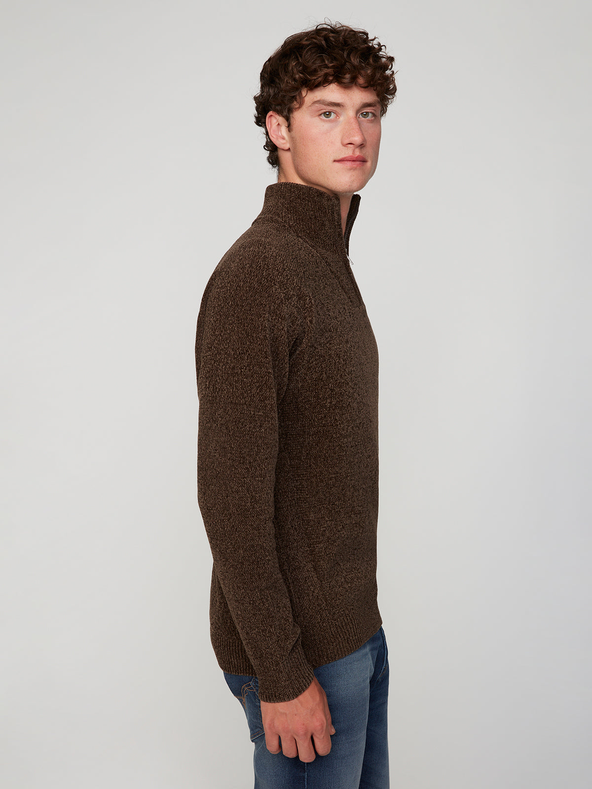 A young man with curly brown hair stands sideways against a plain light background, wearing blue jeans and a PROJEK RAW MENS CHENILLE MOCK NECK SWEATER, looking at the camera.