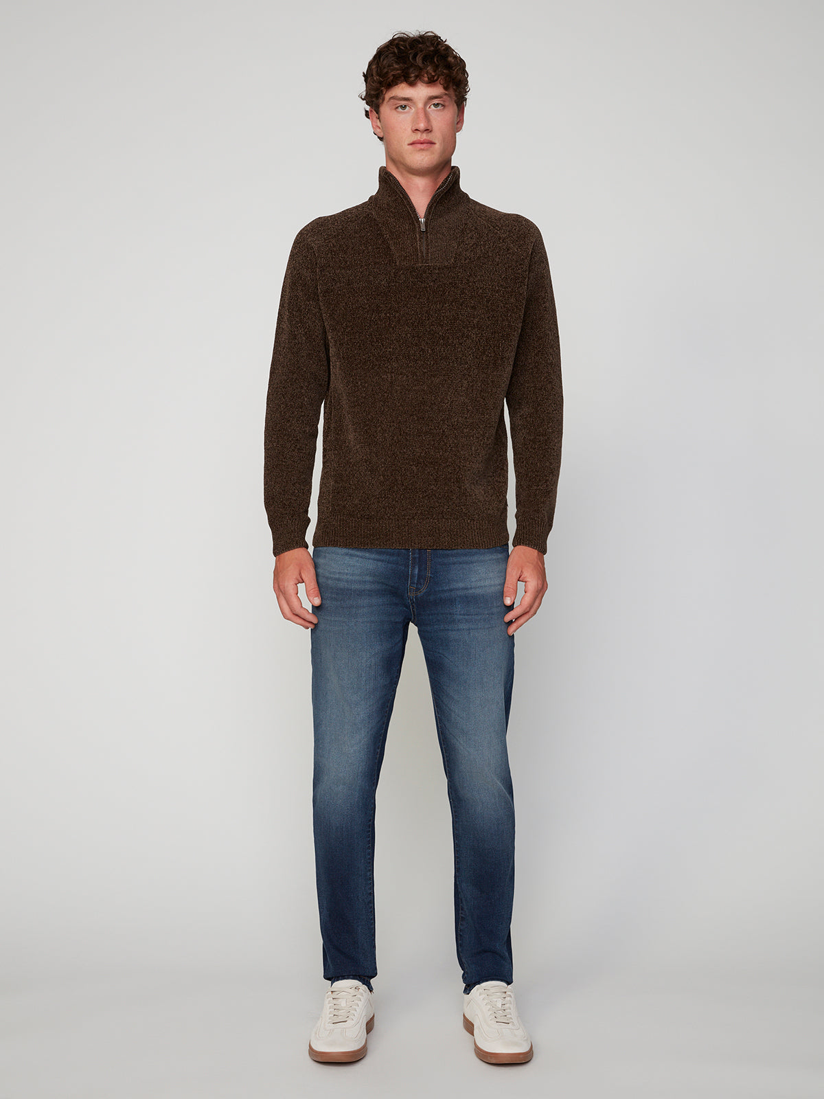 A man stands against a plain light gray background, wearing a brown PROJEK RAW MENS CHENILLE MOCK NECK SWEATER, blue jeans, and white sneakers. He faces forward with his arms relaxed at his sides.