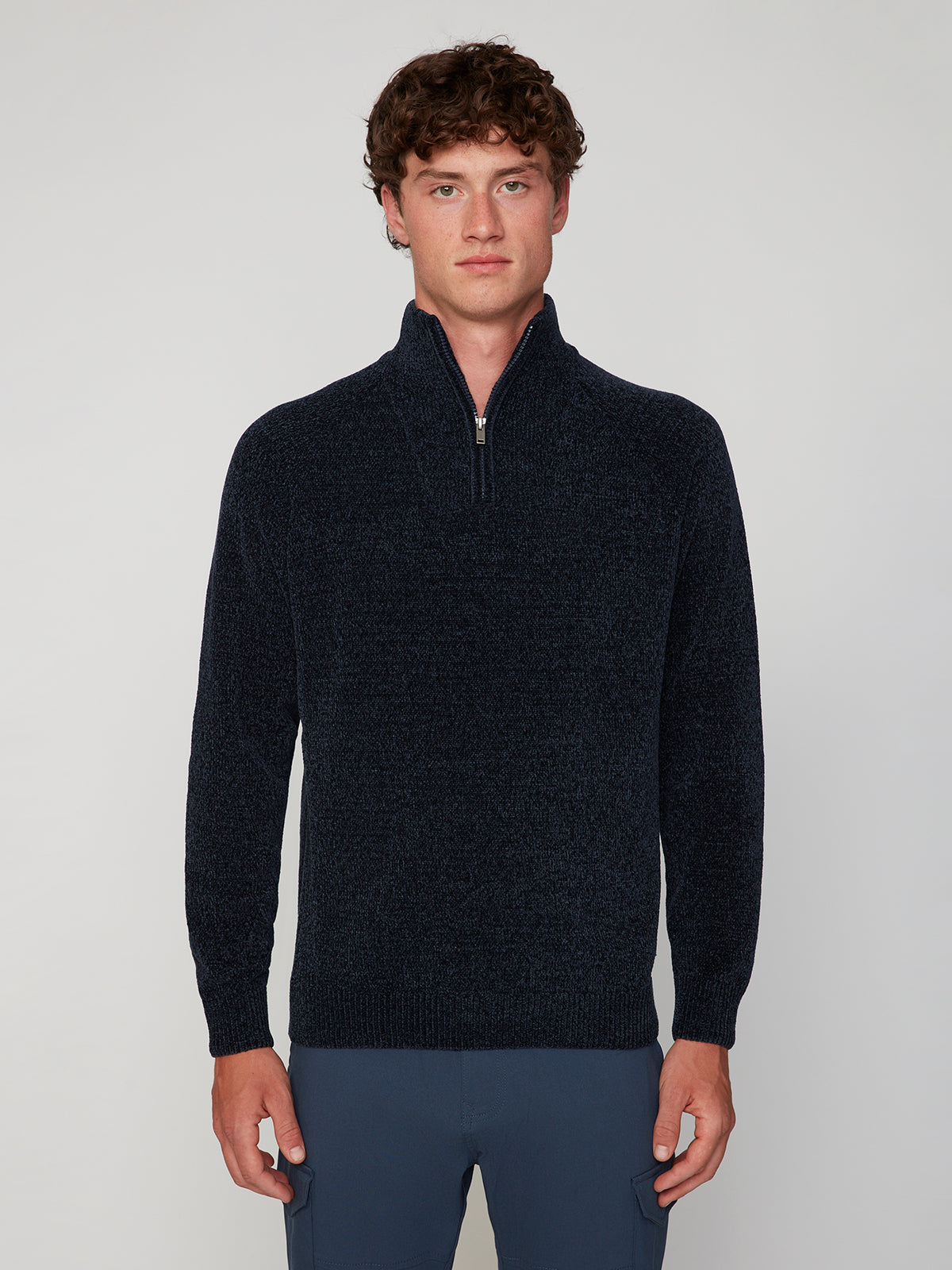 A young man with curly brown hair wears the PROJEK RAW MENS CHENILLE MOCK NECK SWEATER in dark navy and blue pants, standing against a plain light gray background.