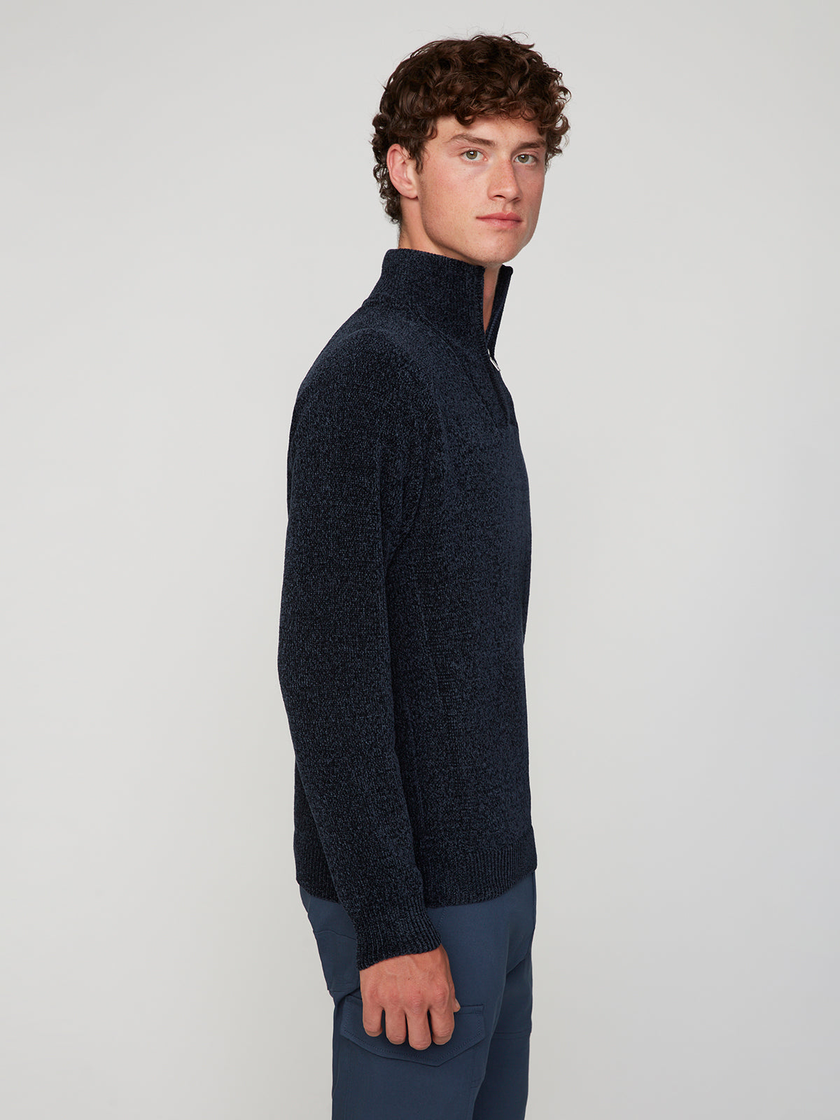 A young man with curly brown hair wears a PROJEK RAW MENS CHENILLE MOCK NECK SWEATER and blue pants, standing sideways and looking at the camera against a plain light background.