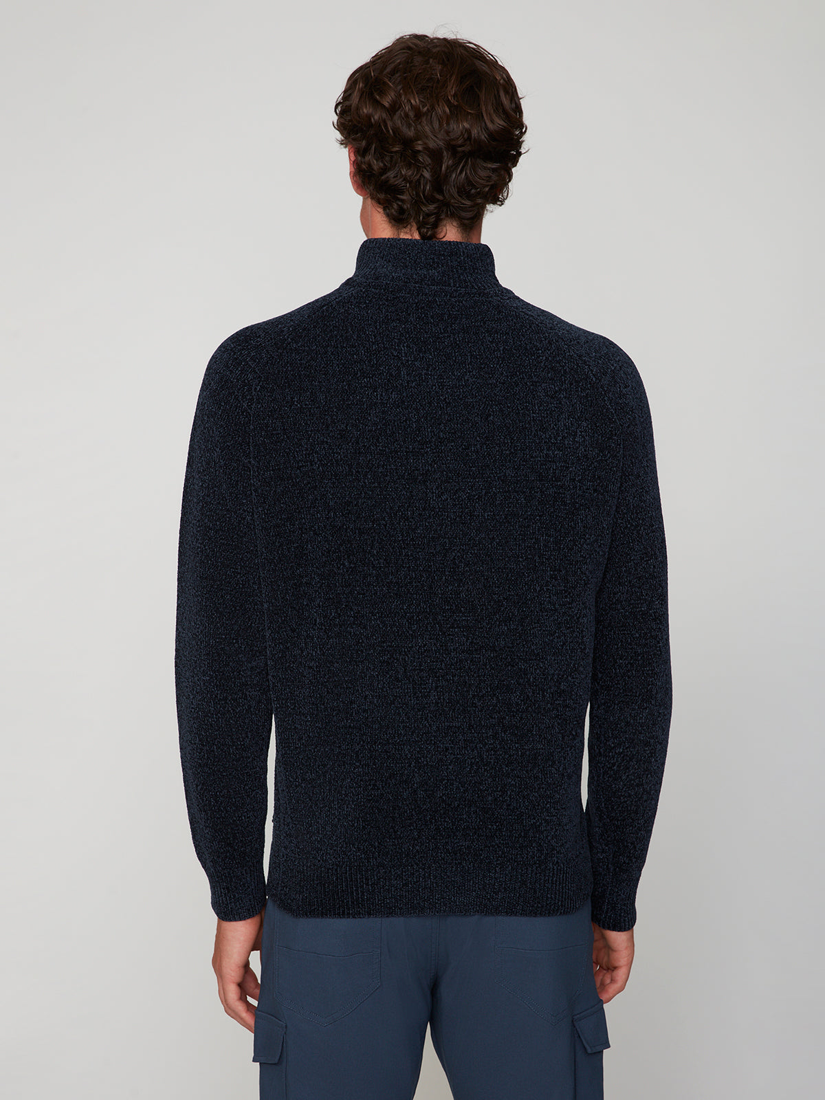 Shown from the back, a person with short, curly brown hair wears the PROJEK RAW MENS CHENILLE MOCK NECK SWEATER in dark navy with blue cargo pants against a plain light gray background.