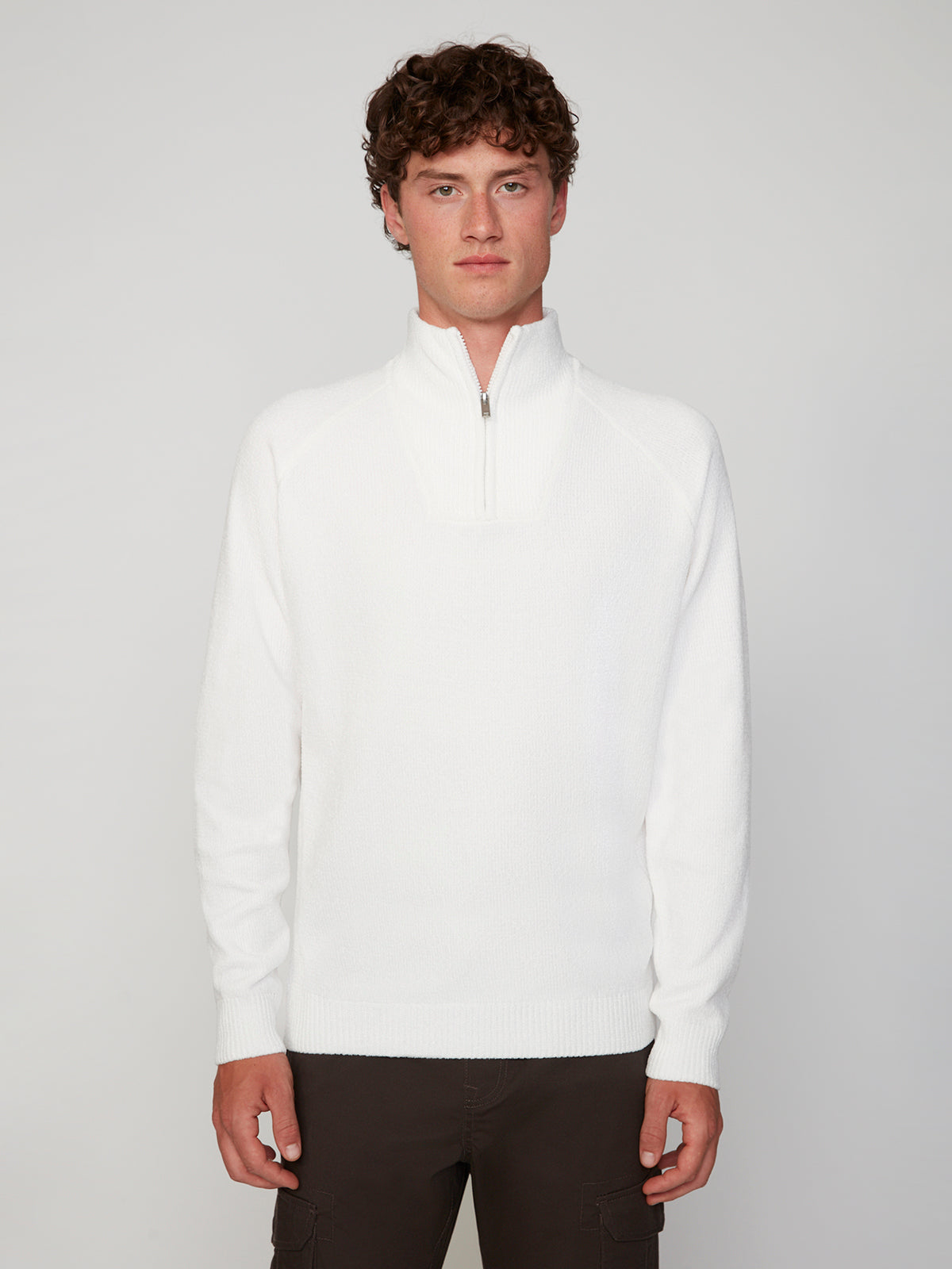 A young man with curly brown hair wears a white PROJEK RAW MENS CHENILLE MOCK NECK SWEATER and dark pants, standing against a plain light gray background, looking directly at the camera with a neutral expression.