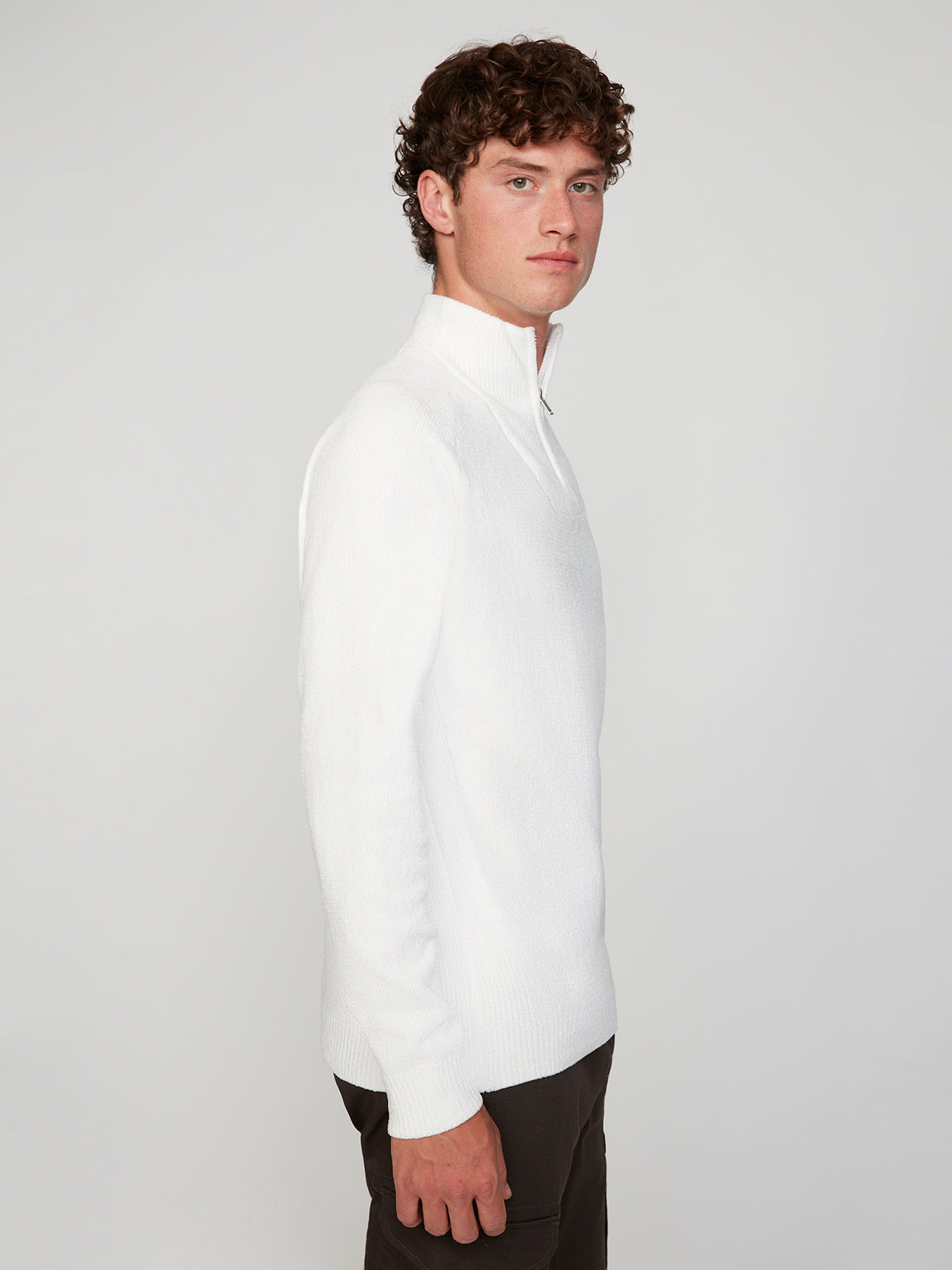 A young man with curly brown hair stands in profile against a plain light background, wearing the PROJEK RAW MENS CHENILLE MOCK NECK SWEATER and dark pants, gazing ahead with a neutral expression.