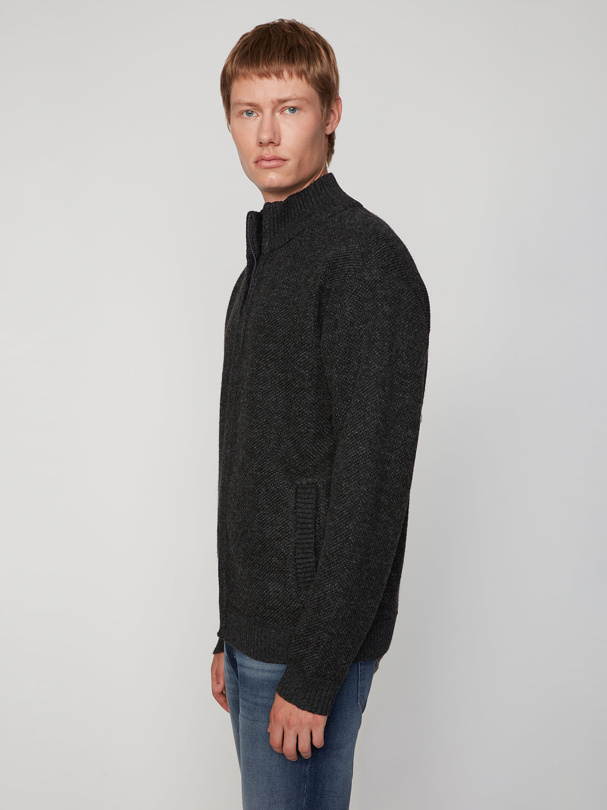 A man with short light brown hair models the PROJEK RAW MENS FULL ZIPPED KNIT SWEATER BONDED POLAR FLEECE in dark gray with blue jeans, standing slightly to the side against a plain light background.