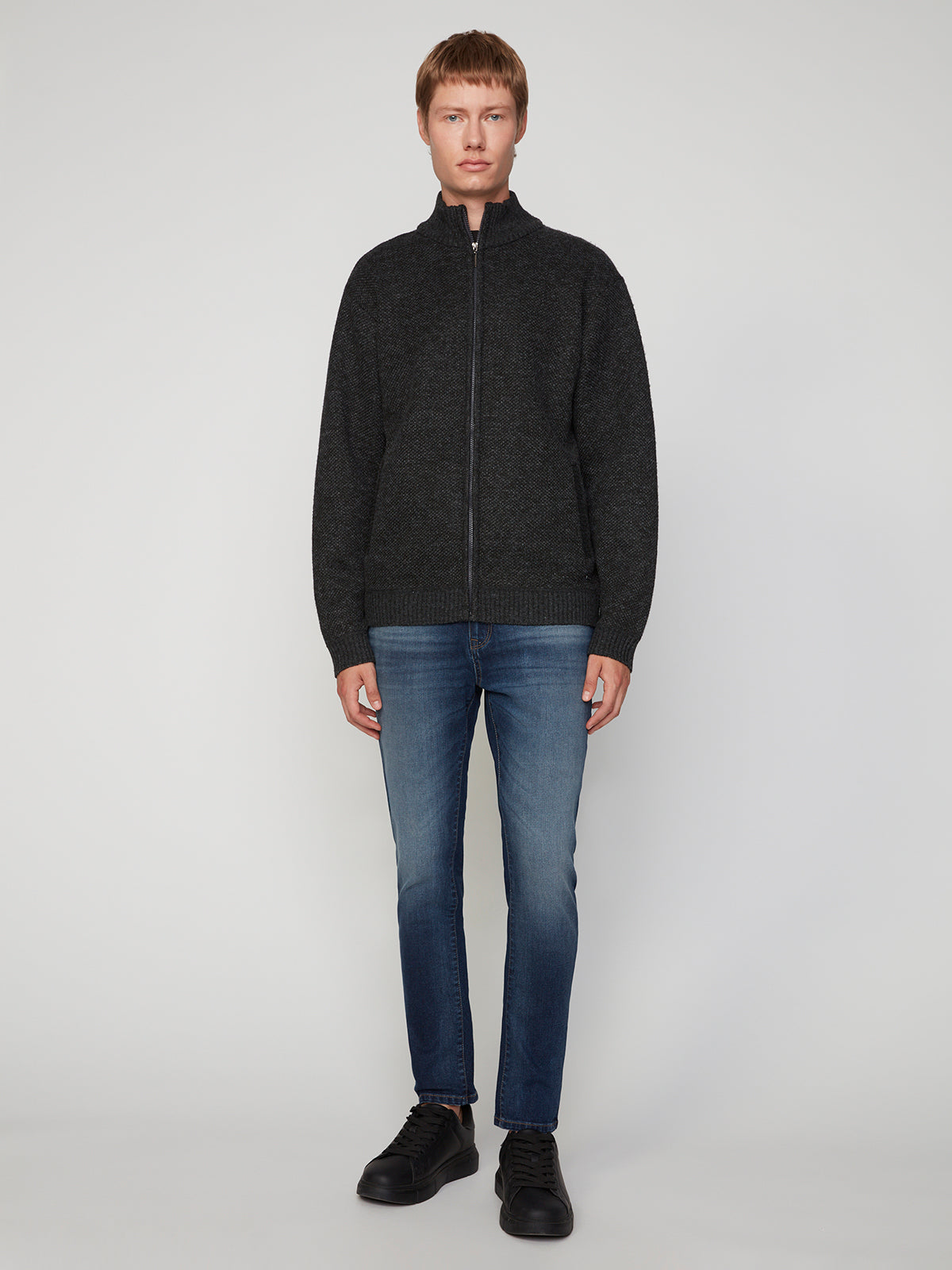 A person with short hair faces forward, wearing PROJEK RAW MENS FULL ZIPPED KNIT SWEATER BONDED POLAR FLEECE, blue jeans, and black shoes against a plain light gray background.