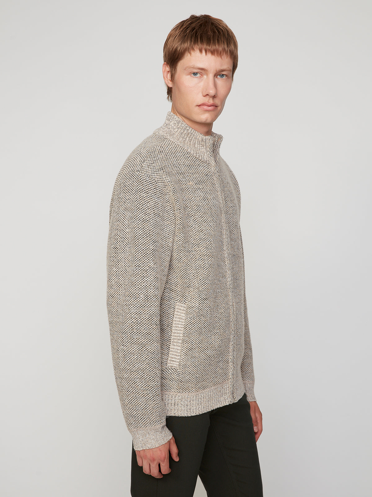 A young man with light brown hair wears the PROJEK RAW MENS FULL ZIPPED KNIT SWEATER BONDED POLAR FLEECE in textured light gray with front pockets and black pants, standing against a white background and looking at the camera.